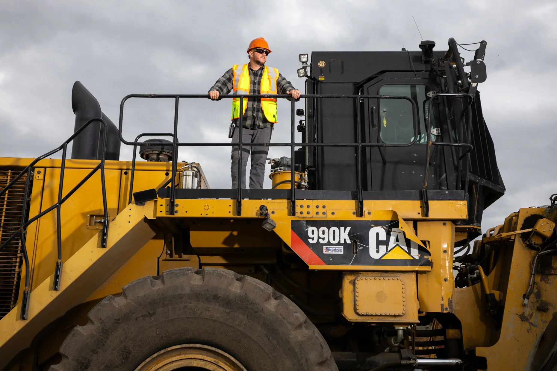 A construction worker stands on a yellow heavy-duty earth-moving machine, wearing a hard hat, sunglasses, and a safety vest, with a cloudy sky in the background.
