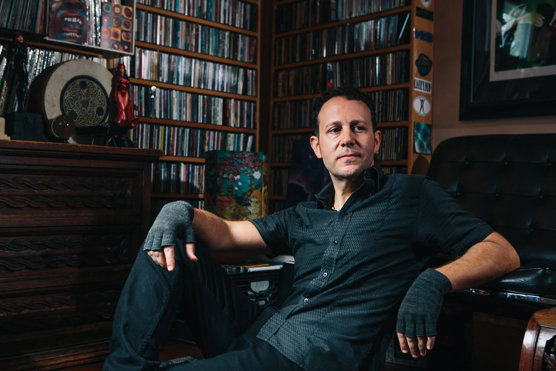 A man with short dark hair and a slight beard, wearing a black shirt with rolled-up sleeves, sitting on a black sofa in a room filled with shelves of books and CDs, with artistic decorations and artworks in the background.