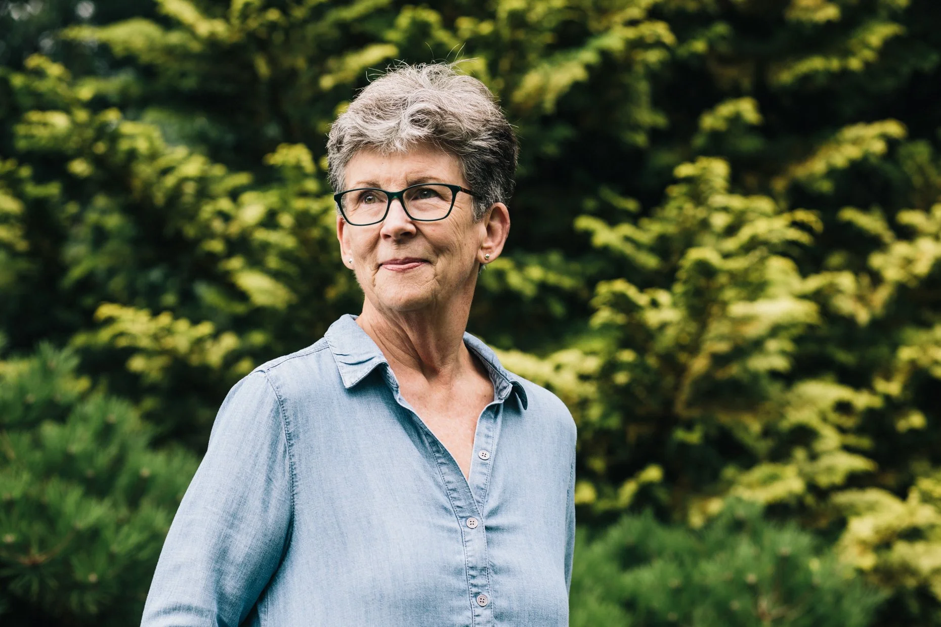 An elderly woman with short gray hair, wearing glasses and a light blue denim shirt, stands outdoors in front of green bushes and trees, looking pensively to her left.