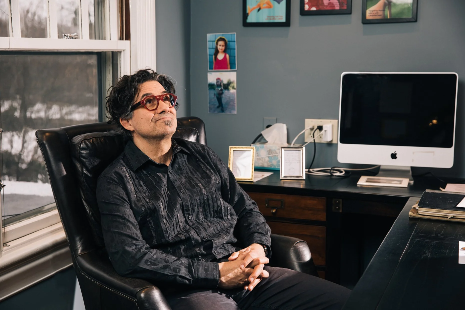 A man with dark curly hair, wearing red glasses and a black patterned shirt, sits in a black leather office chair with his hands clasped, looking upwards in a modern office space near a window with snowy trees outside. On the wall behind him are framed artwork and a photograph of a smiling girl. A computer monitor and desk items are visible on the right.
