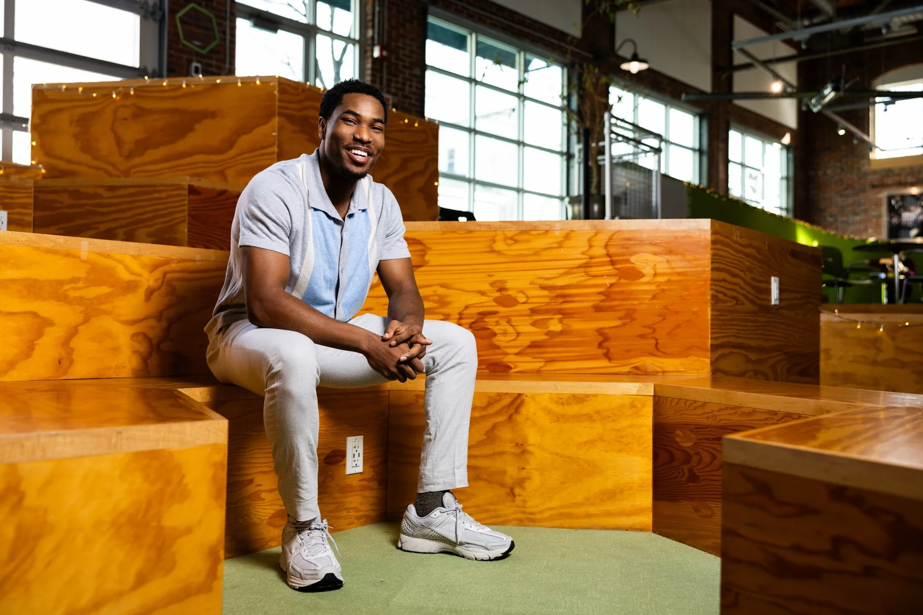 A smiling man with dark skin and short curly hair, wearing a light blue polo shirt, beige pants, and white sneakers, sitting on wooden bleacher-style seats inside a modern, well-lit indoor space with large windows, brick walls, and green accents.