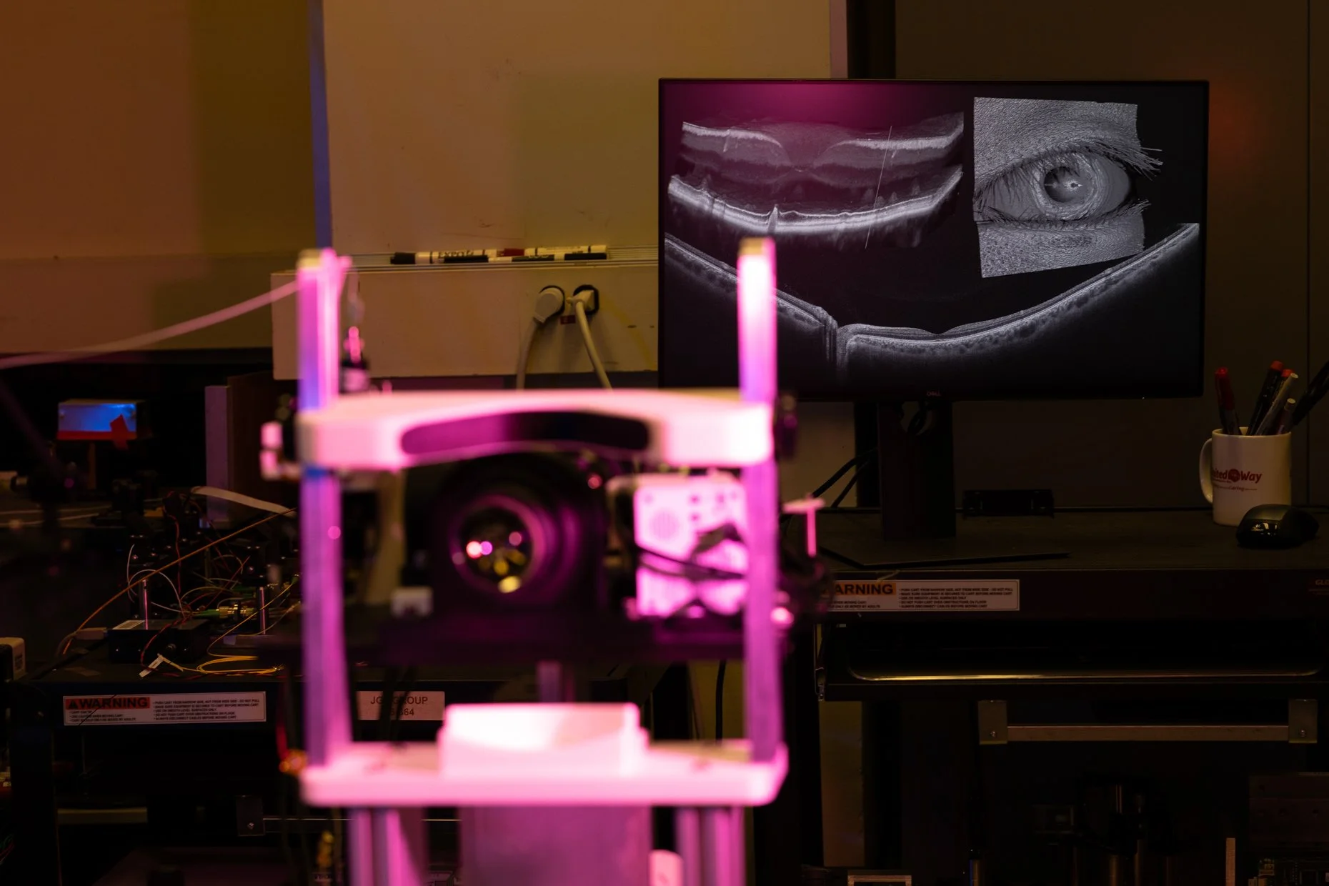 A workspace with a 3D printer in the foreground, emitting pink light. In the background, a monitor displays black and white images of an eye and an MRI scan. There are various wires, tools, and equipment on the work surface.