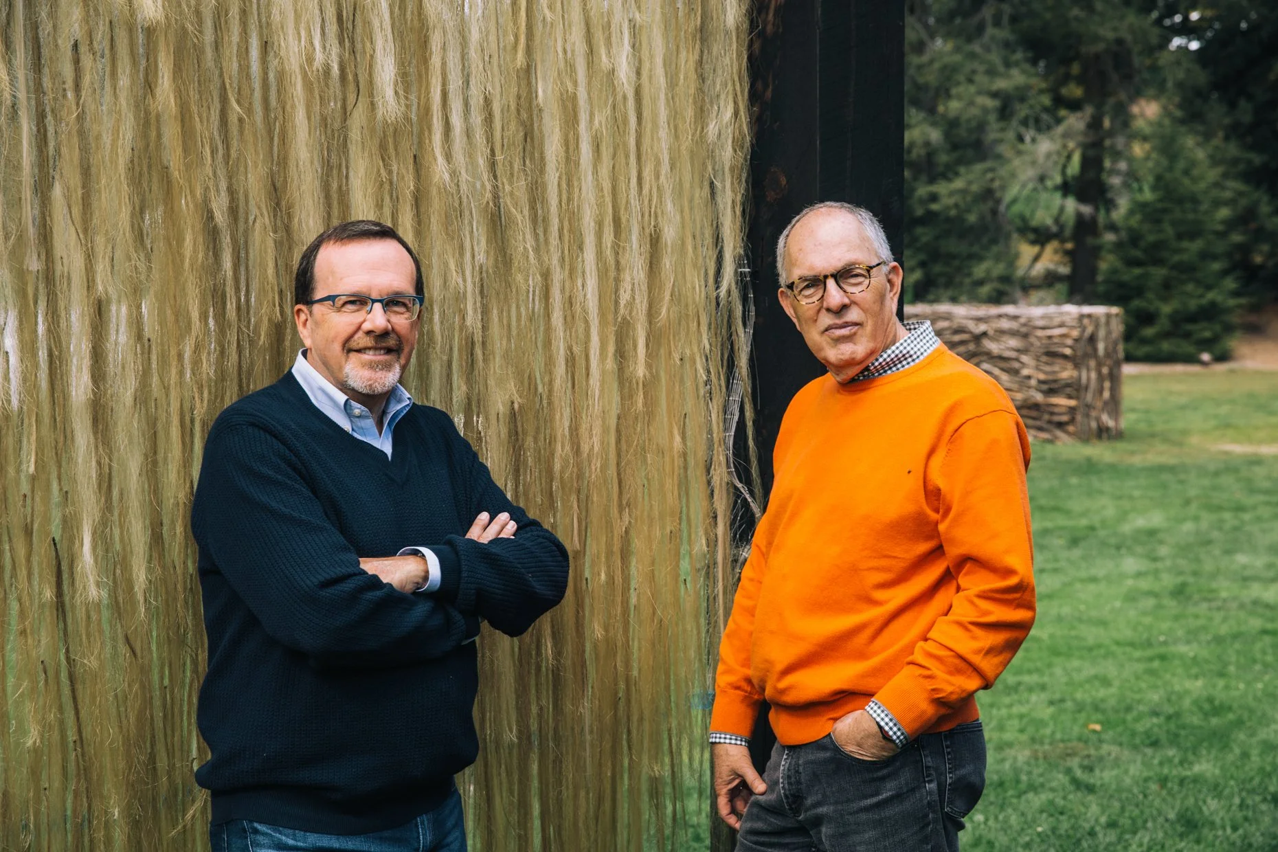 Two middle-aged men standing outdoors near a large hanging installation of light-colored vertical fibers. One man is wearing a dark sweater and glasses, with his arms crossed. The other man is in an orange sweater, also wearing glasses, with his hand in his pocket. A natural park setting is visible in the background.