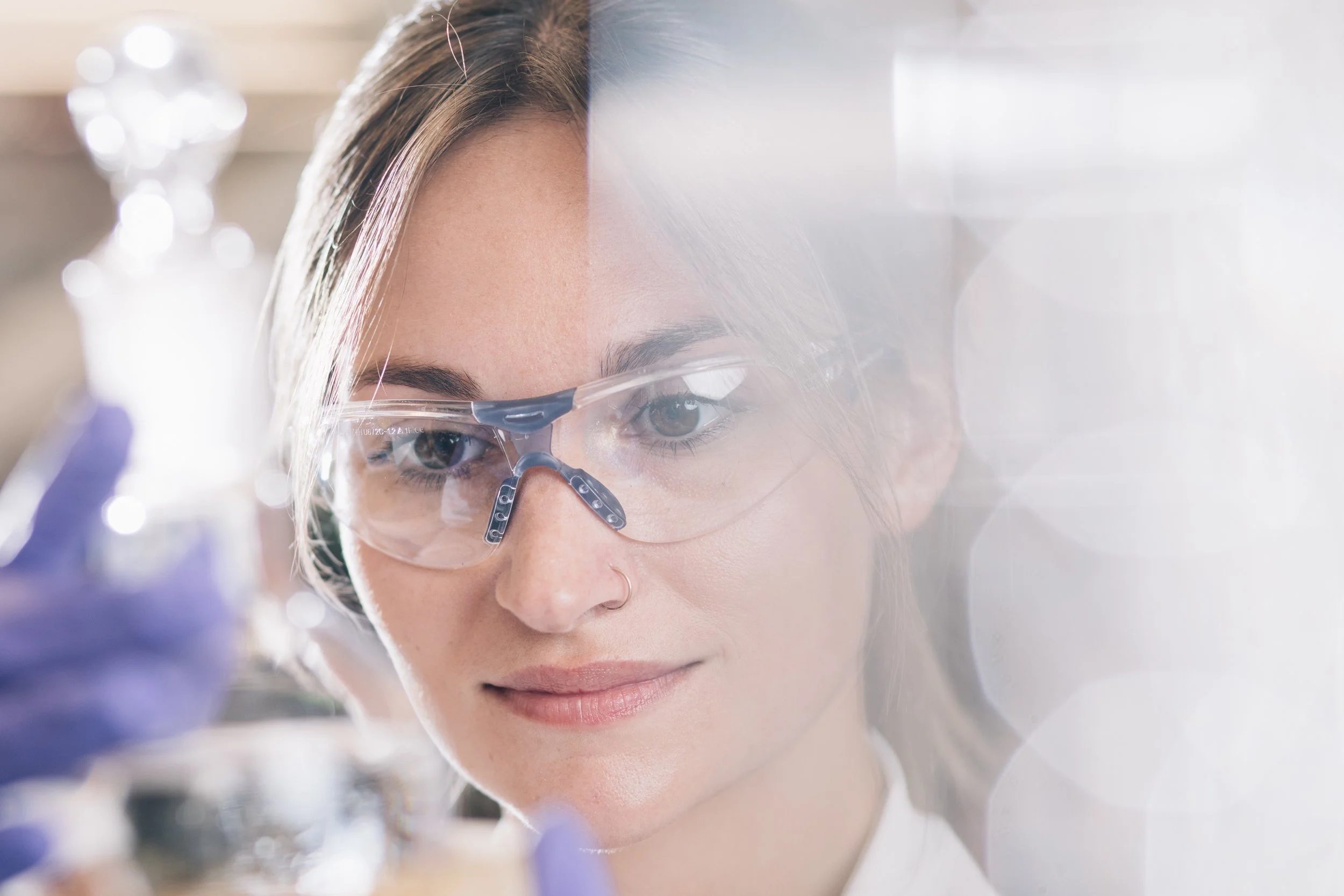 A woman with light skin wearing safety glasses is working in a laboratory or scientific setting, with focus on her face and eyes.