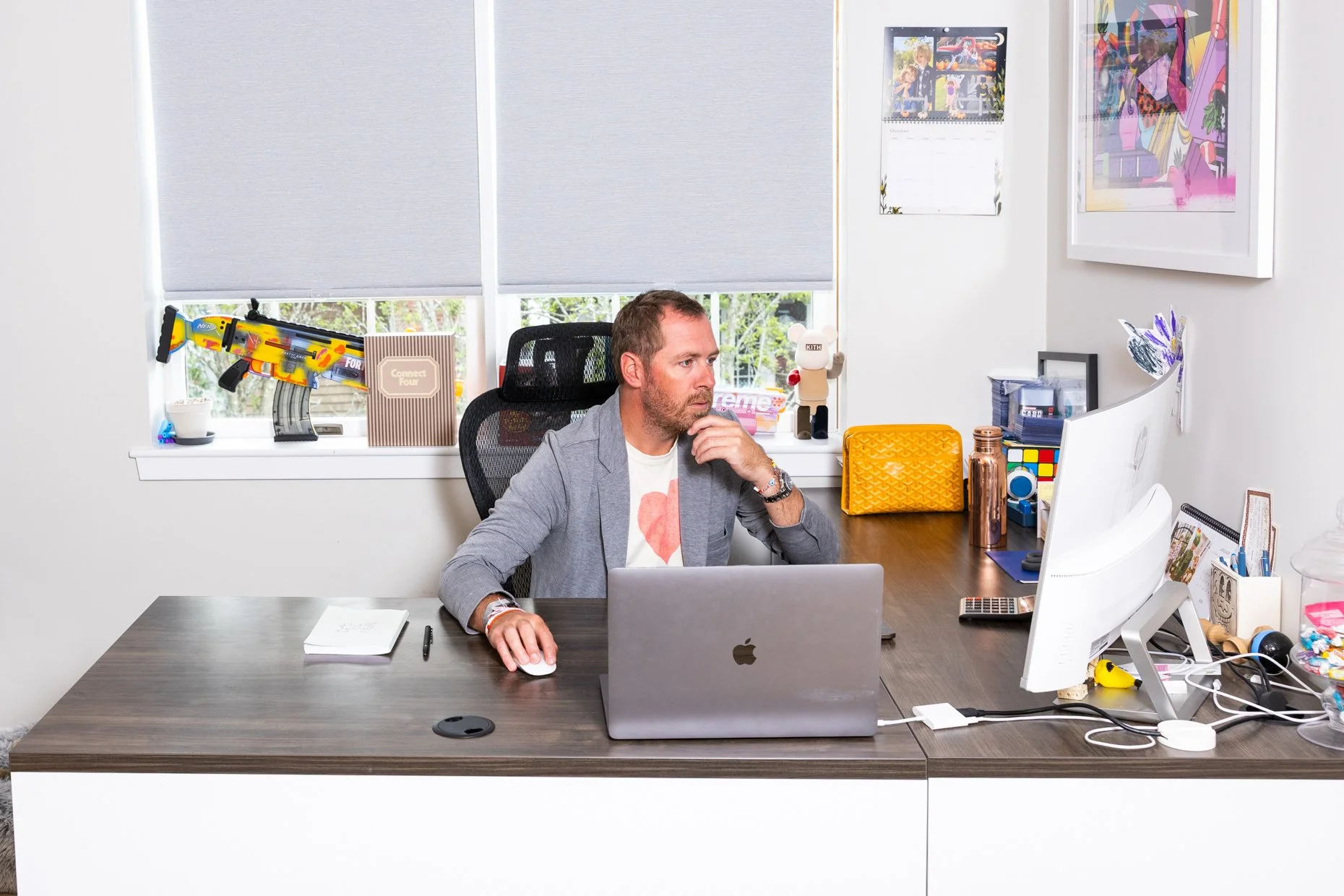 Office space with a man sitting at a desk working on a laptop. The desk is cluttered with various items, and there's a window with blinds behind him. A toy gun is visible on the windowsill.