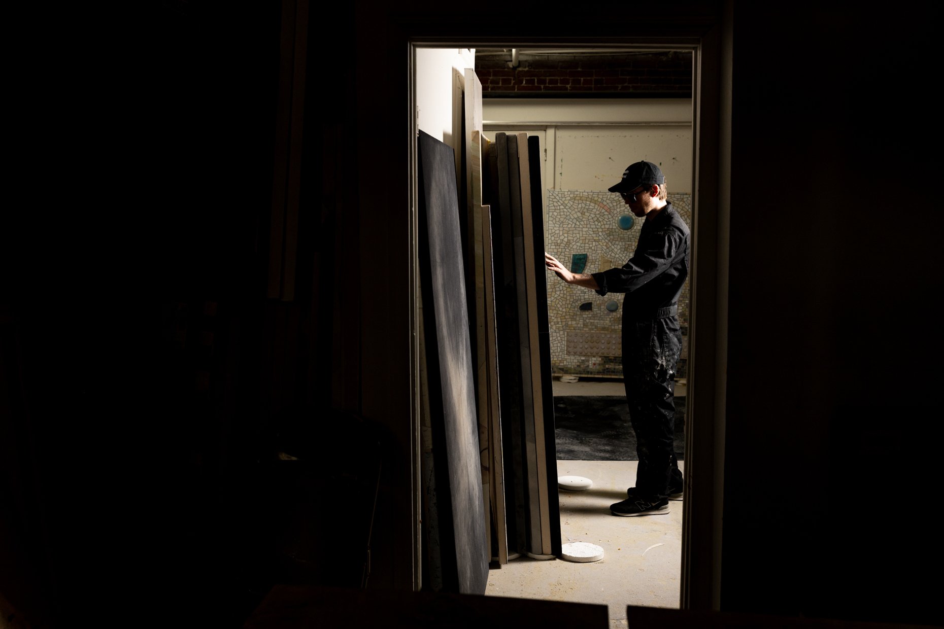A person wearing a black cap, sunglasses, and a black jumpsuit is looking at or working with pieces of large glass or acrylic panels in a workshop or studio with construction or art supplies around.