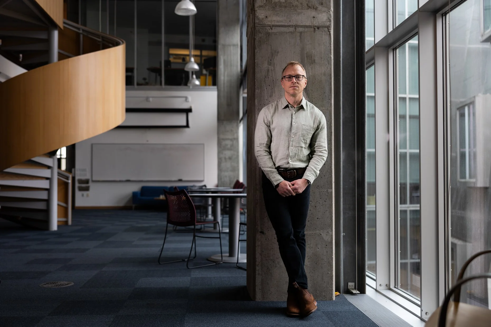 Man with glasses and a light gray shirt standing by a large window in a modern office space with concrete walls, wooden staircase, and office furniture.