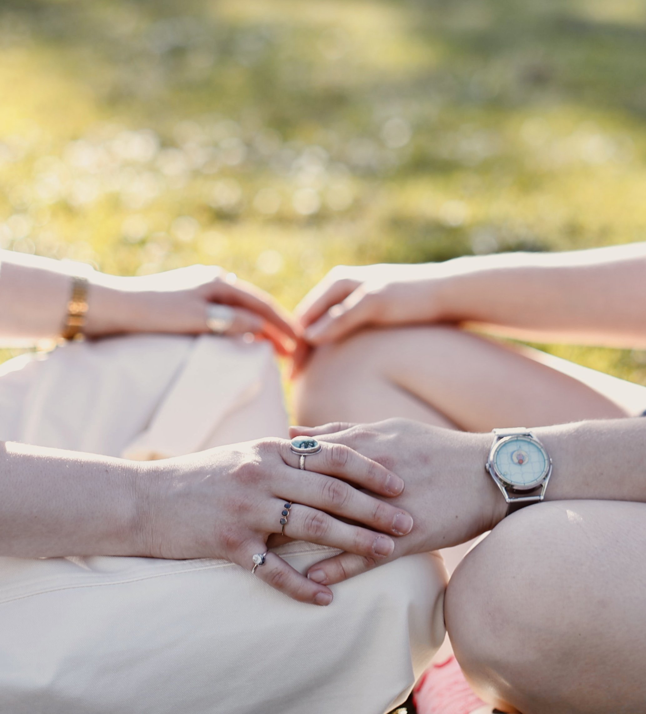 Close-up of several hands with rings, placed on a person's stomach, with a blurred background of nature or outdoors.