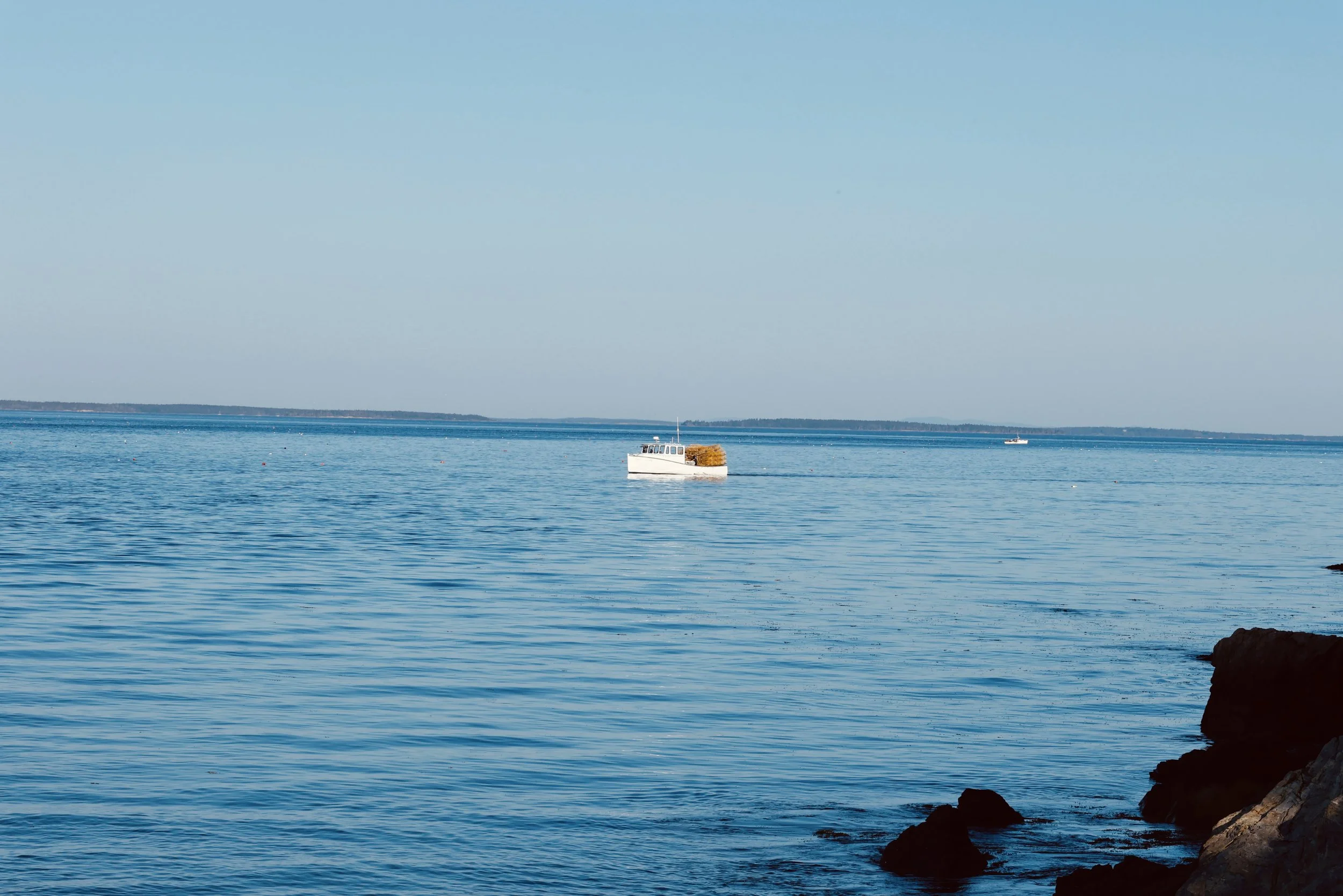 A calm body of water with a small boat carrying hay, and another boat in the distance, under a clear blue sky.