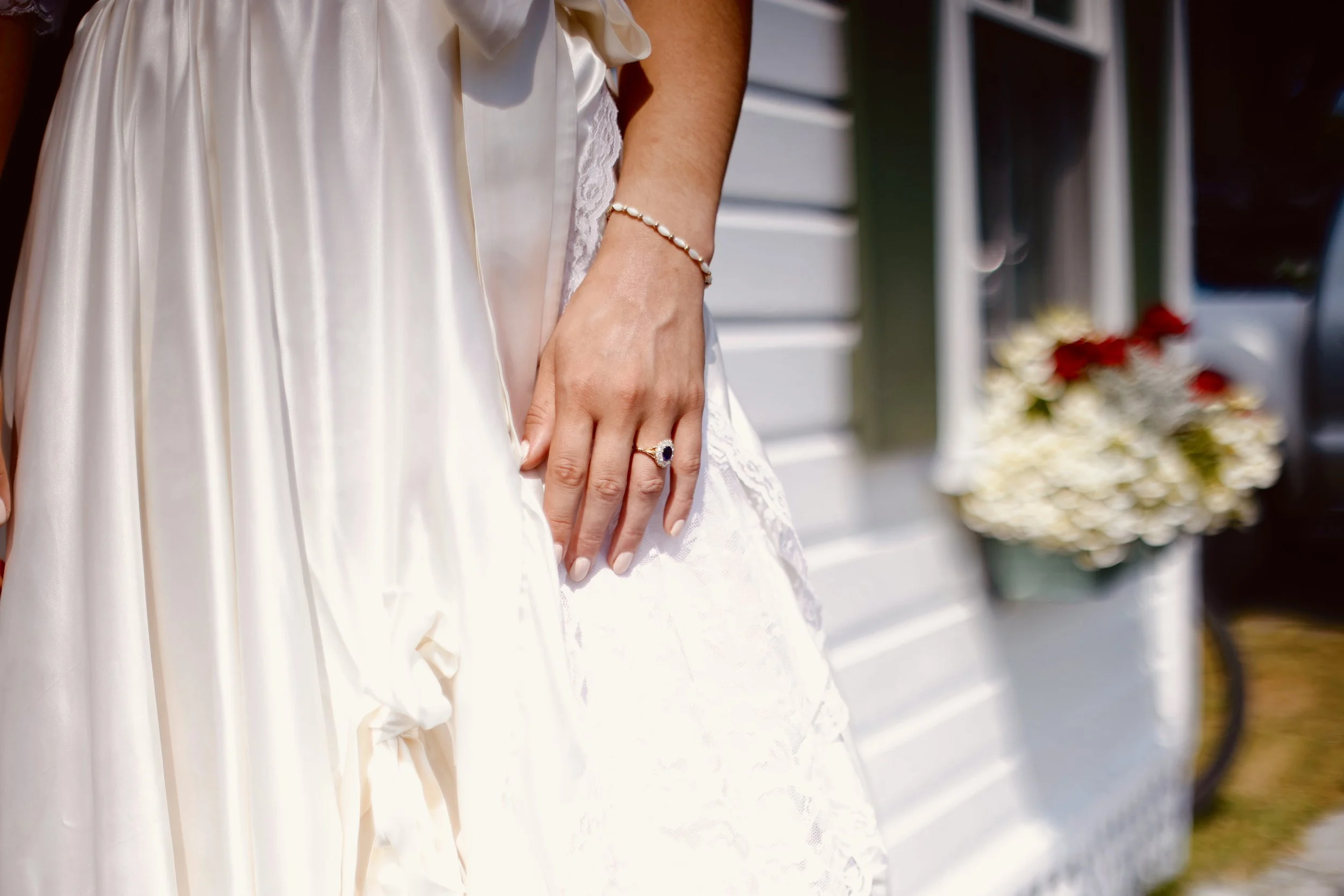 Close-up of a woman wearing a white satin dress with lace details, jewelry including a pearl bracelet and a ring, outdoors near a white fence and flowers.