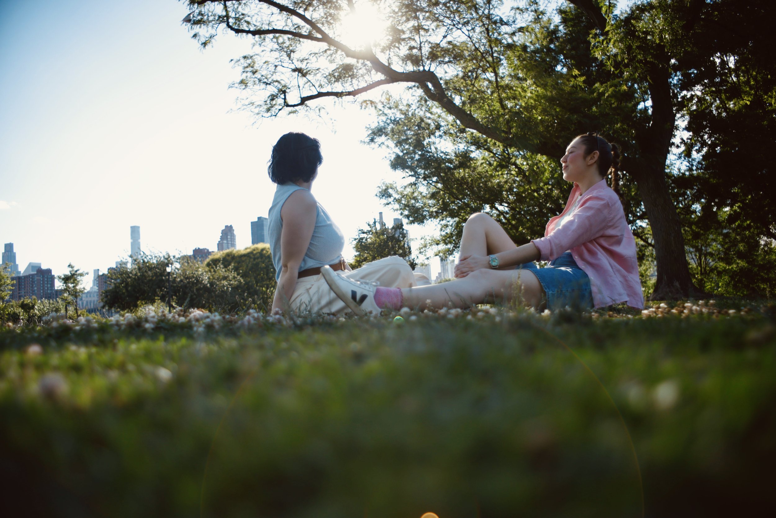 Two women sitting on grass under a large tree in a park, with a city skyline in the background on a sunny day.