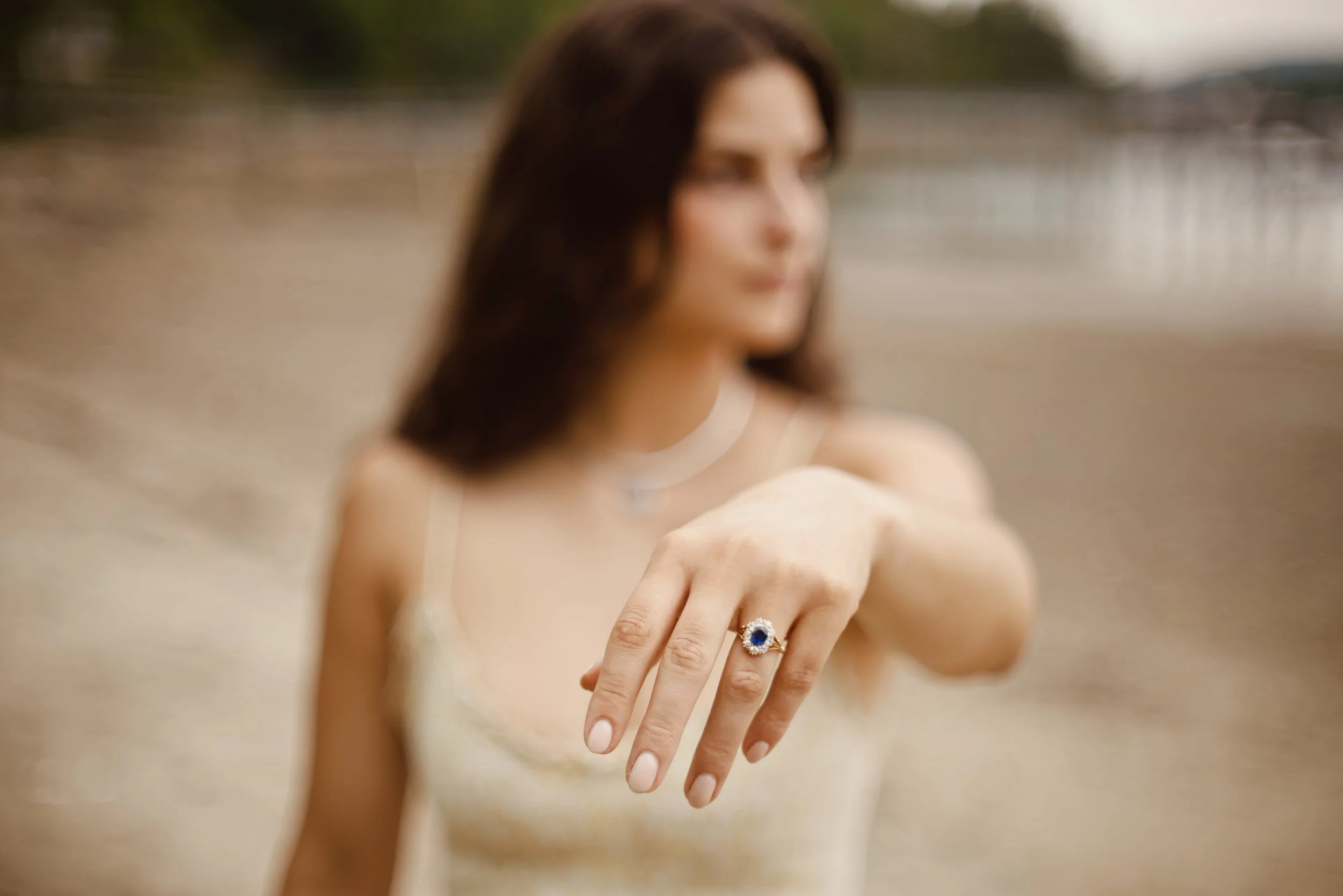 Close-up of a woman's hand showing off a ring with a blue sapphire ring, in focus, blurred background with her face slightly out of focus.