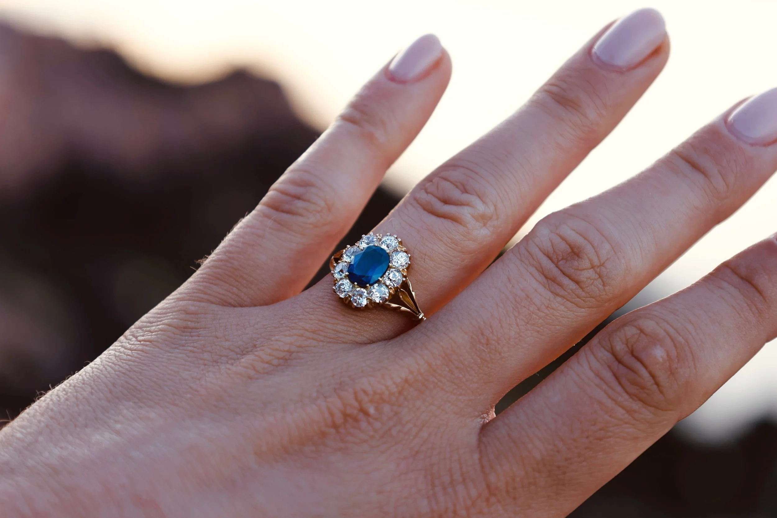 Close-up of a hand wearing a gold ring with a large blue gemstone surrounded by small white diamonds.