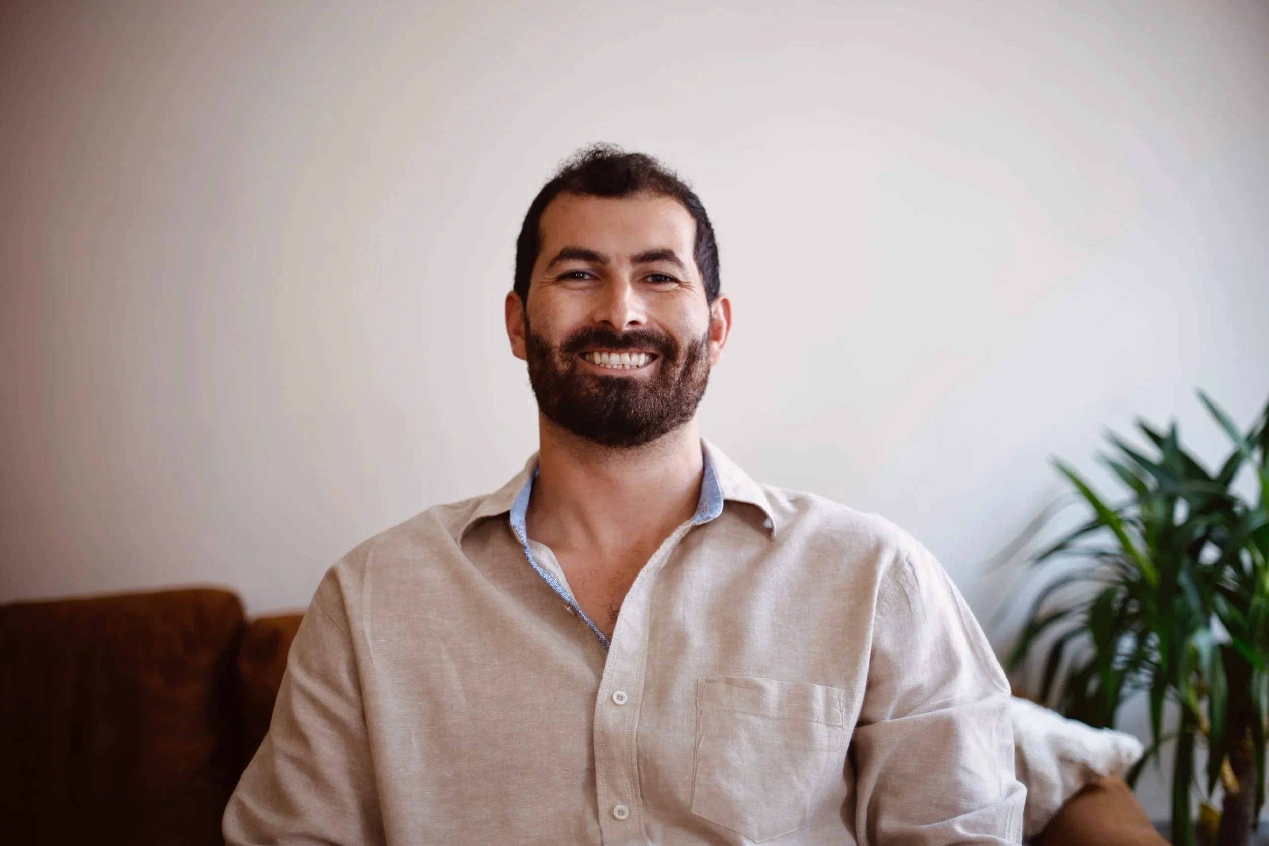 A smiling man with dark hair and a beard, wearing a beige button-up shirt, sitting indoors near a green plant, in a room with a white wall.