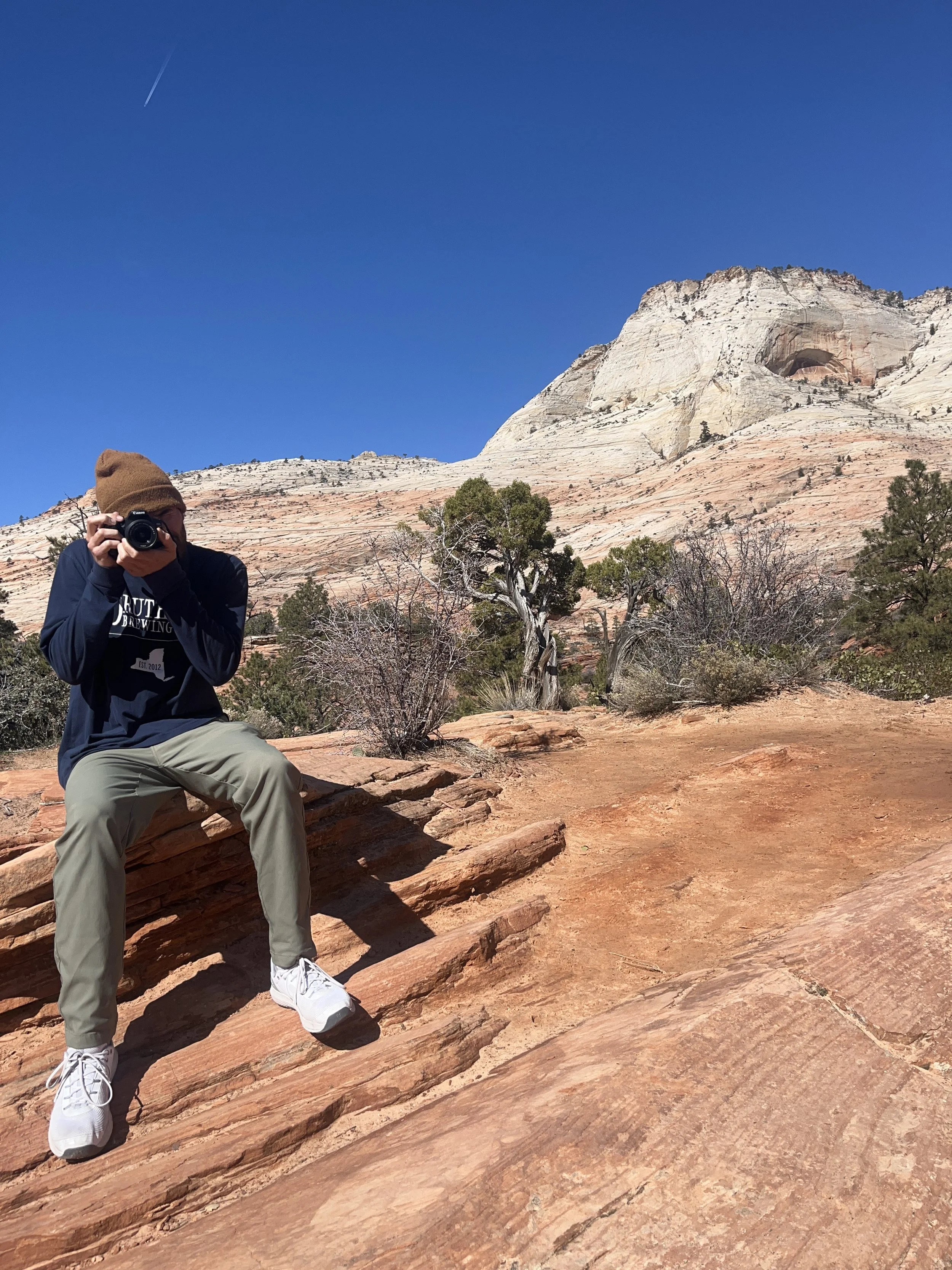 Person wearing brown beanie and dark hoodie taking a photo with a camera in a desert landscape with layered rock formations and sparse trees under a clear blue sky.
