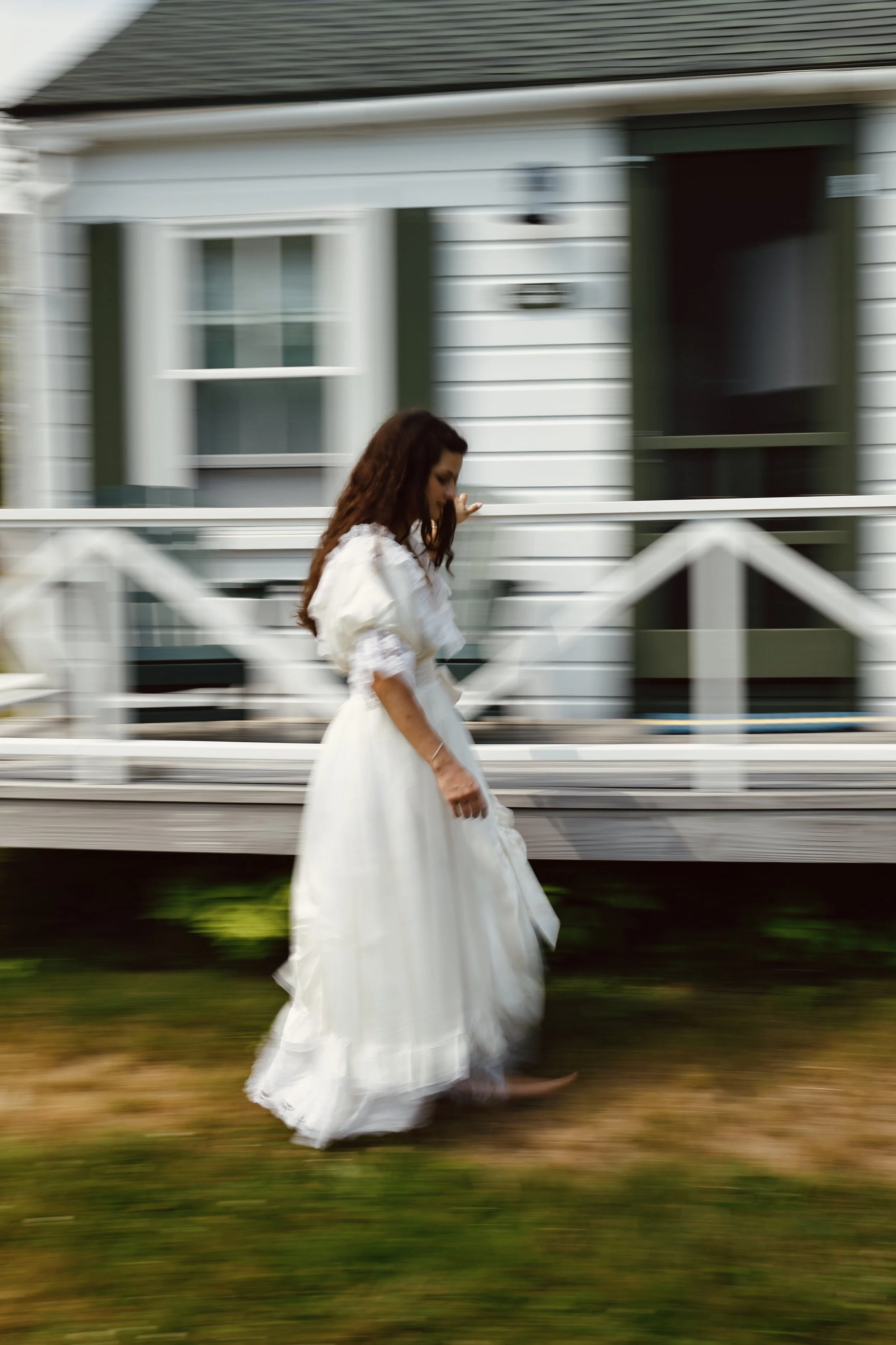 A woman in a white dress walking past a house with white siding and green shutters.