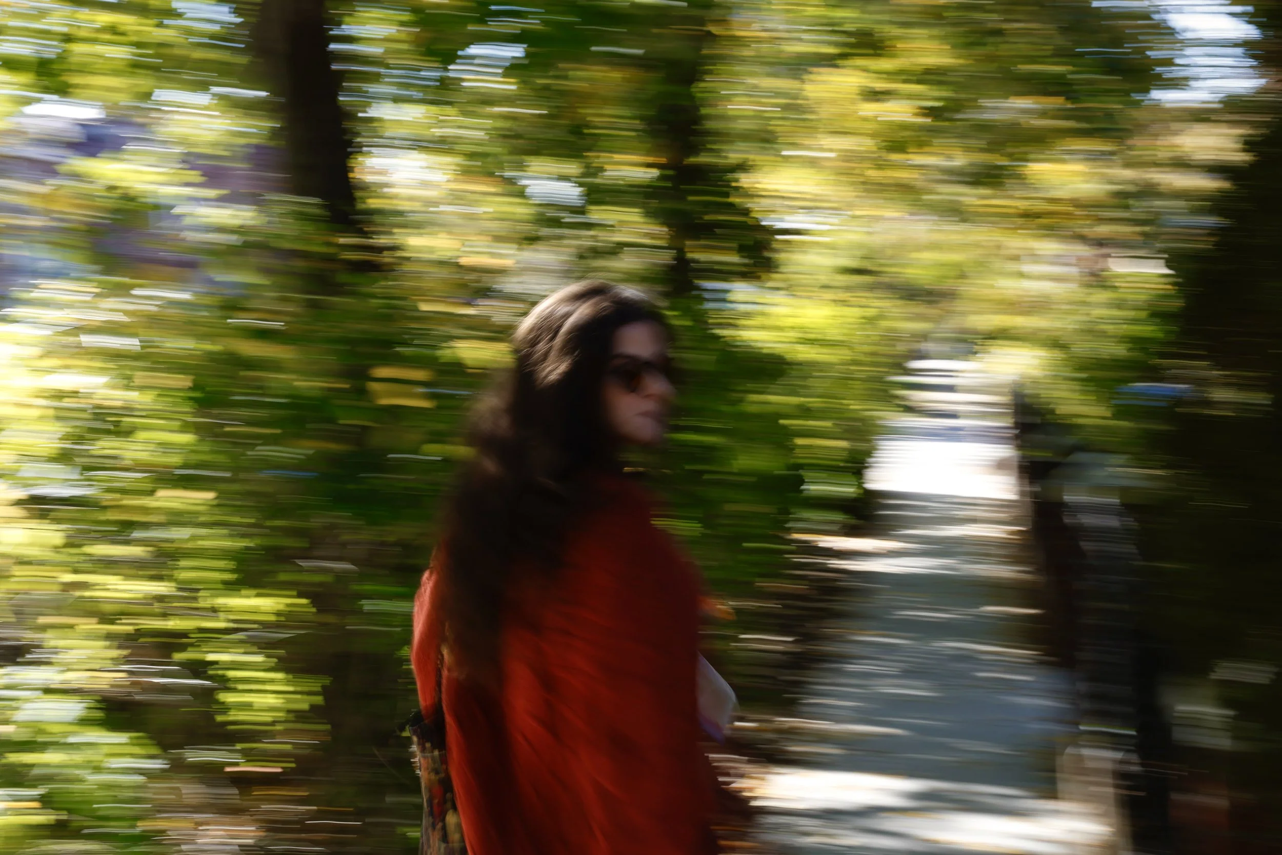 A woman with long dark hair wearing sunglasses and a red coat walking outdoors among blurred green trees with sunlight filtering through.