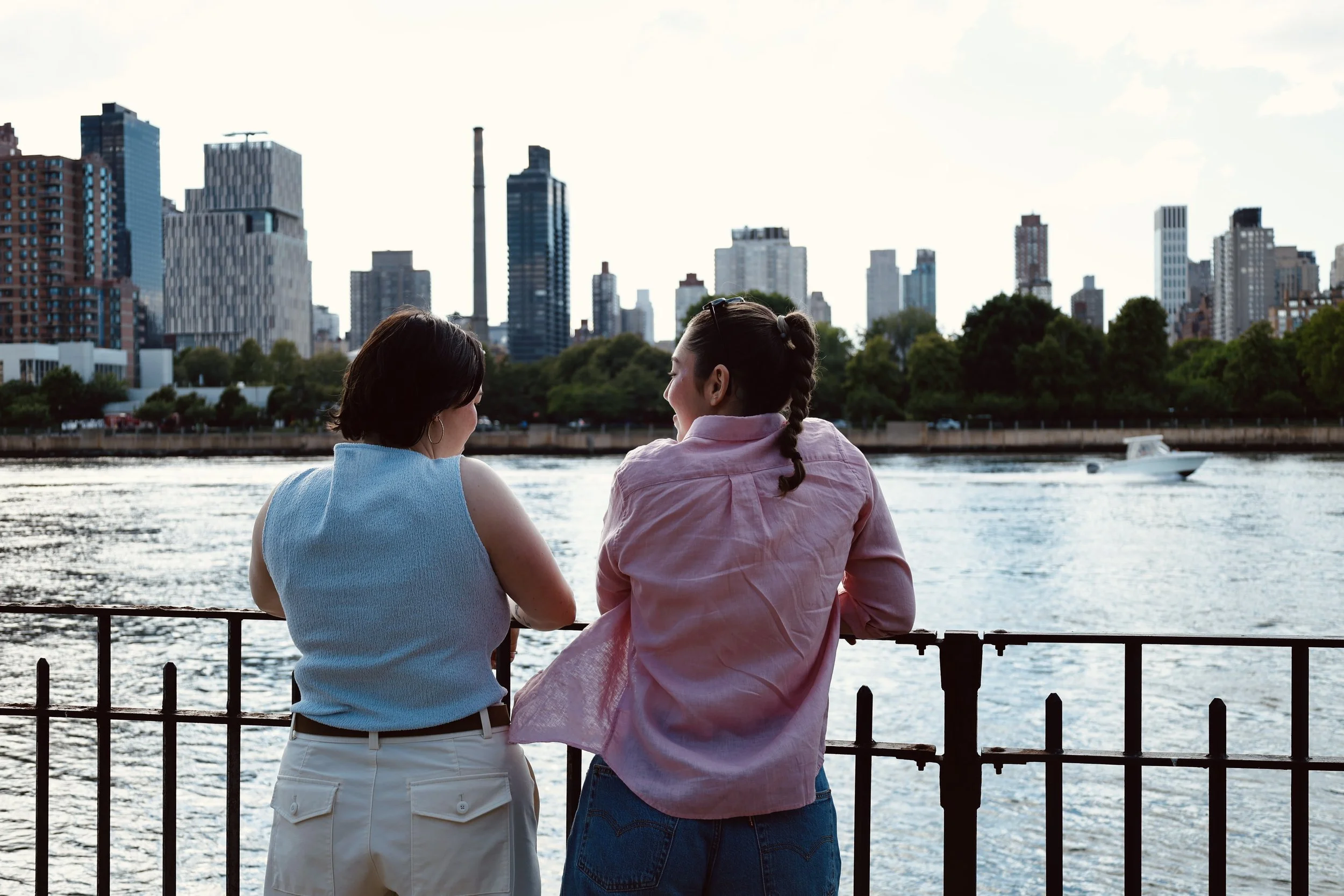 Two women are sitting by a river, looking at the New York City skyline with tall buildings and trees in the background.