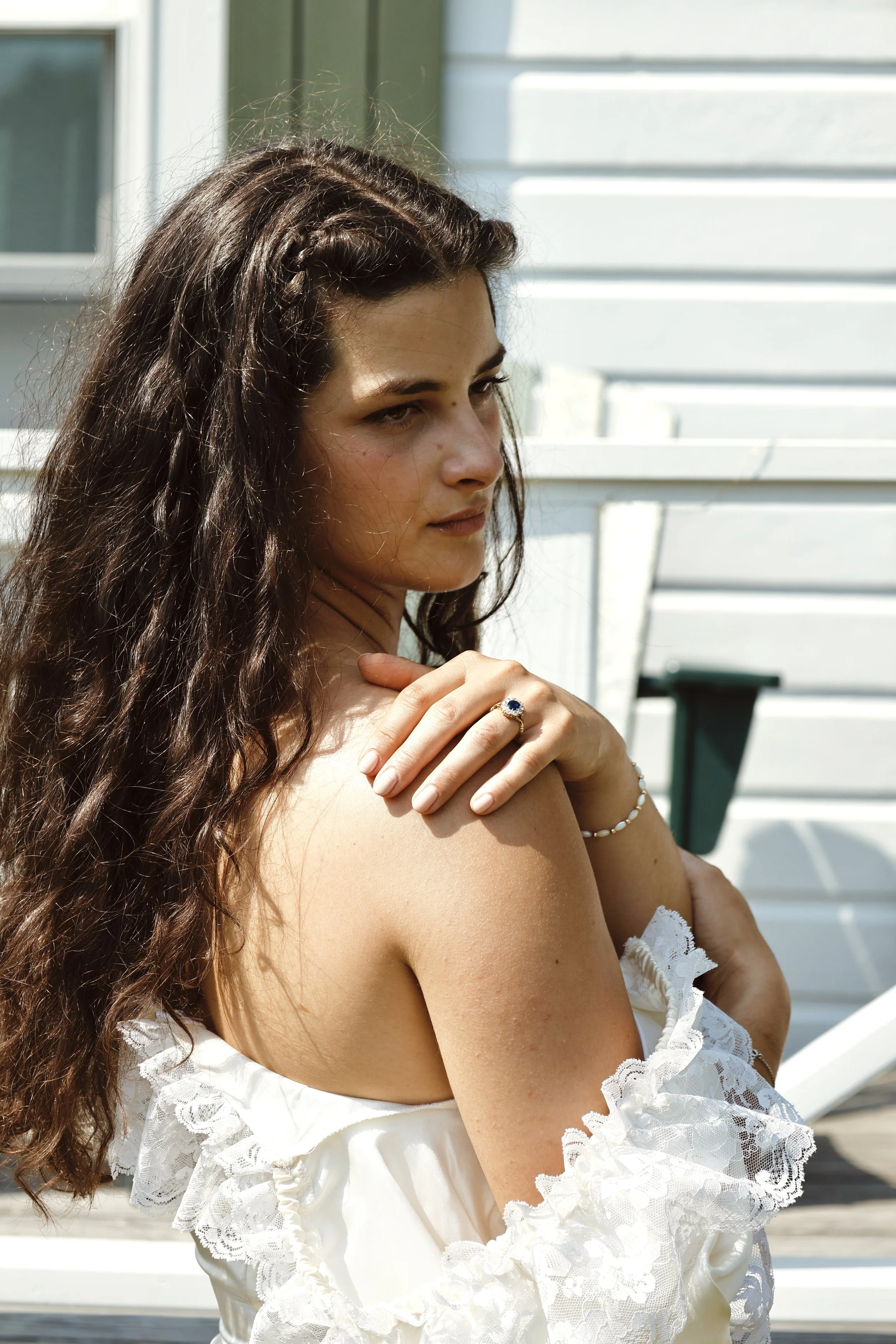 A woman with long, wavy brown hair, wearing a white off-the-shoulder lace dress, standing outdoors near a white bench.