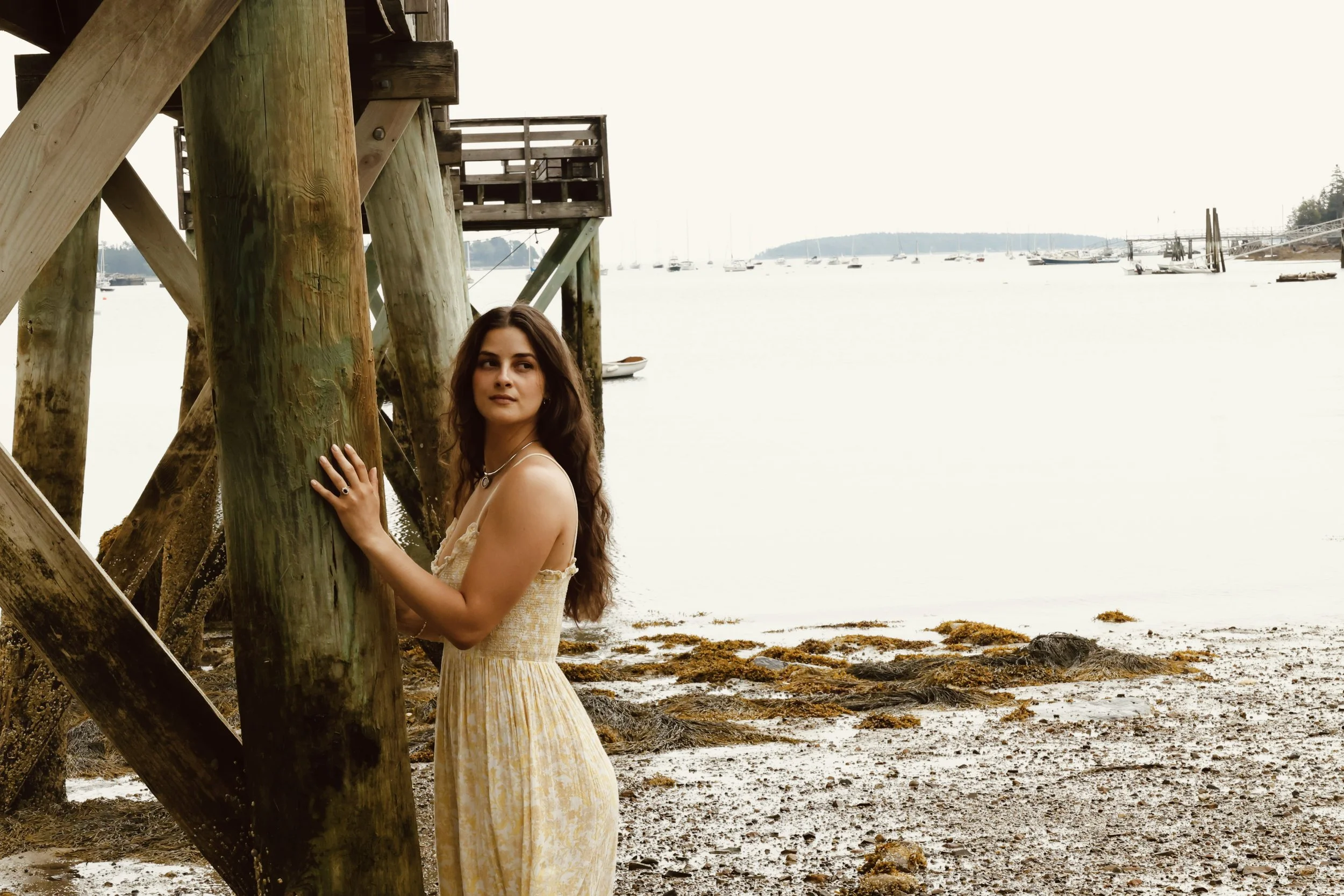 A woman in a yellow dress with brown hair, standing near wooden pilings by a beach with boats and sailboats in the background.