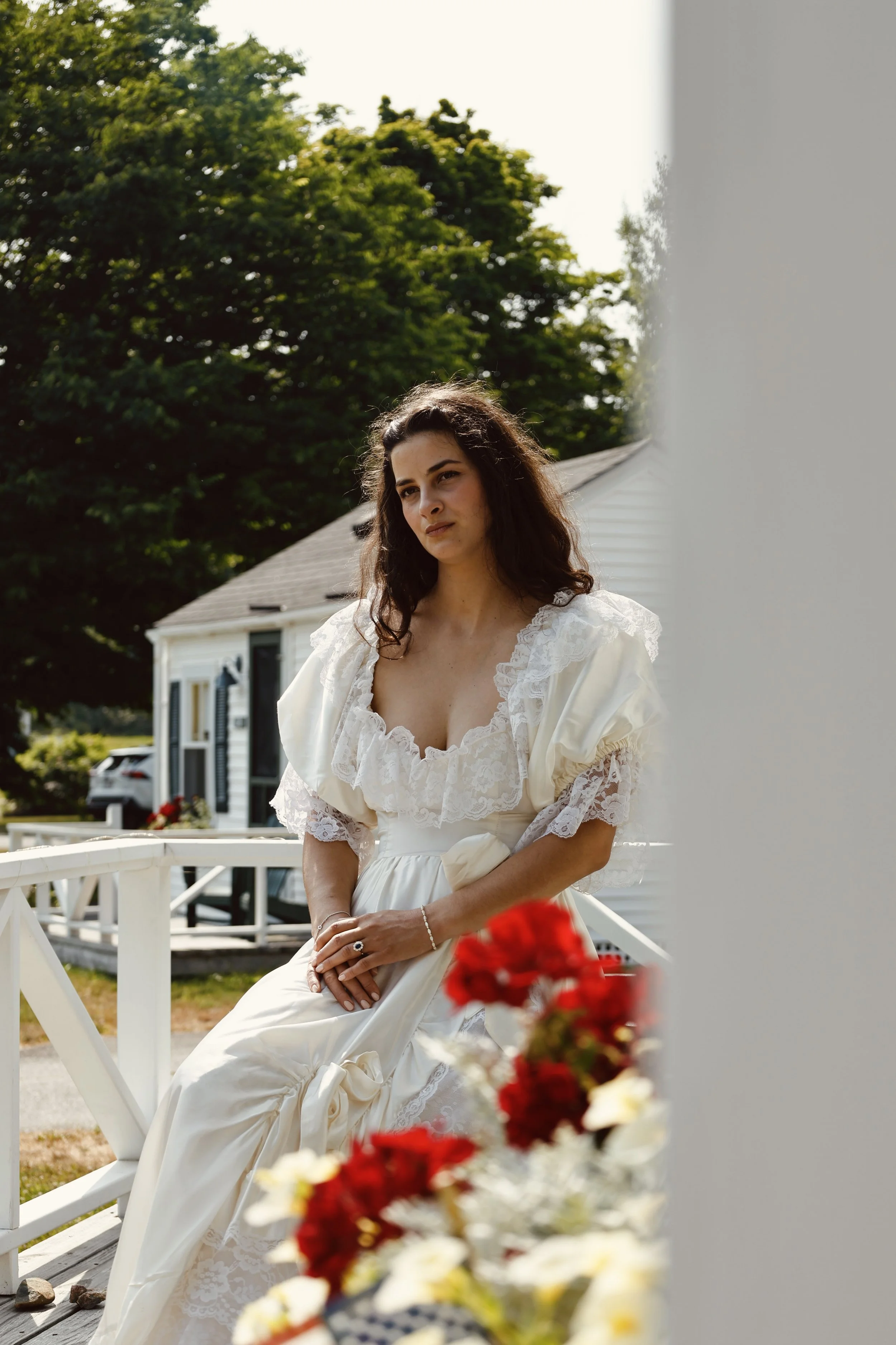 A woman dressed in a vintage white lace dress with puffed sleeves is sitting on a white picket fence outside a house, with green trees in the background. She has dark, wavy hair and is looking to the side. She is holding her hands together on her lap