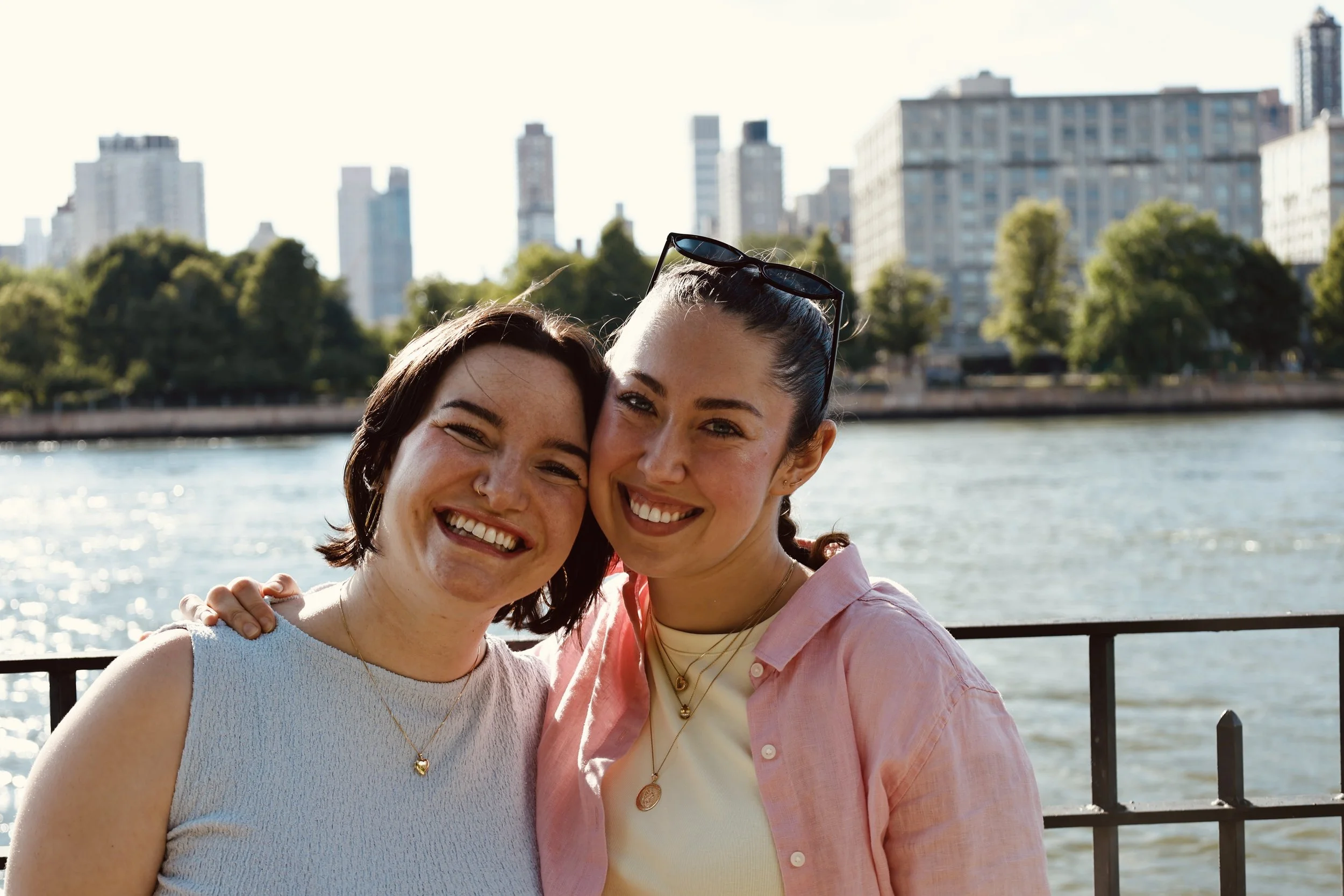 Two smiling women with dark hair and light skin, wearing necklaces, standing close together outdoors near water with a city skyline in the background.