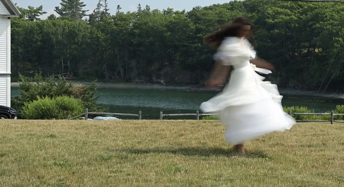 A woman in a flowing white dress twirling outdoors on a grassy area near a body of water with trees in the background.