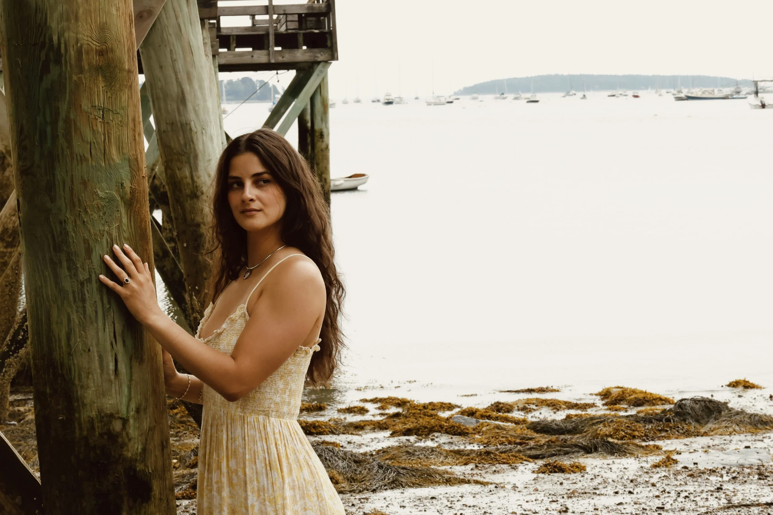 A woman with long, wavy brown hair and light skin is standing beside a wooden pole by a beach, and she is touching the pole with her left hand. She is wearing a light-colored sleeveless dress and a necklace, with a calm expression. The background shows boats anchored on the water and a distant shoreline under a cloudy sky.