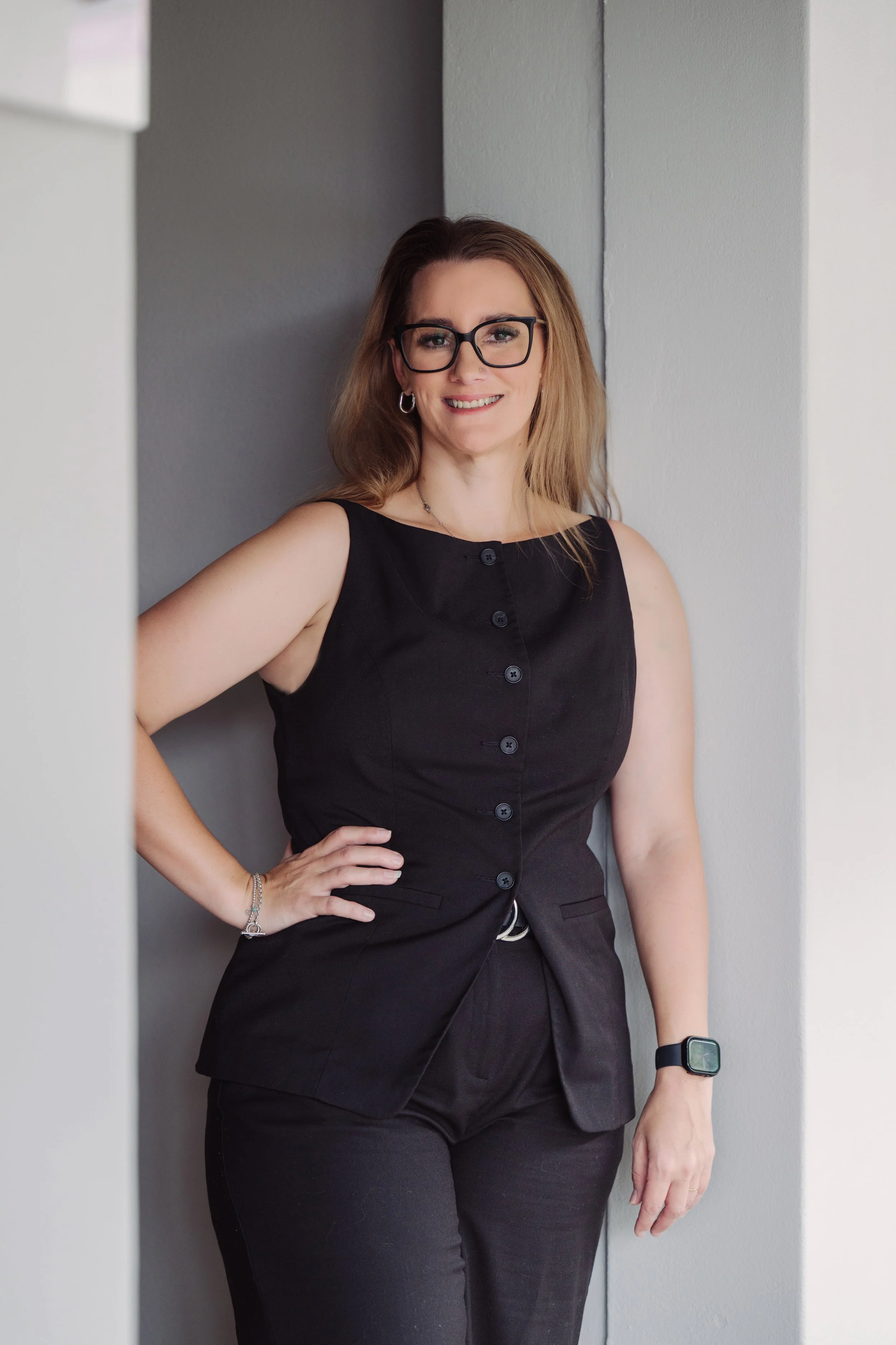 A woman with shoulder-length brown hair, wearing black glasses, a sleeveless black top, and black pants, stands with one hand on her hip, smiling. She is also wearing earrings, a bracelet, and a smartwatch, and is positioned against a gray wall.