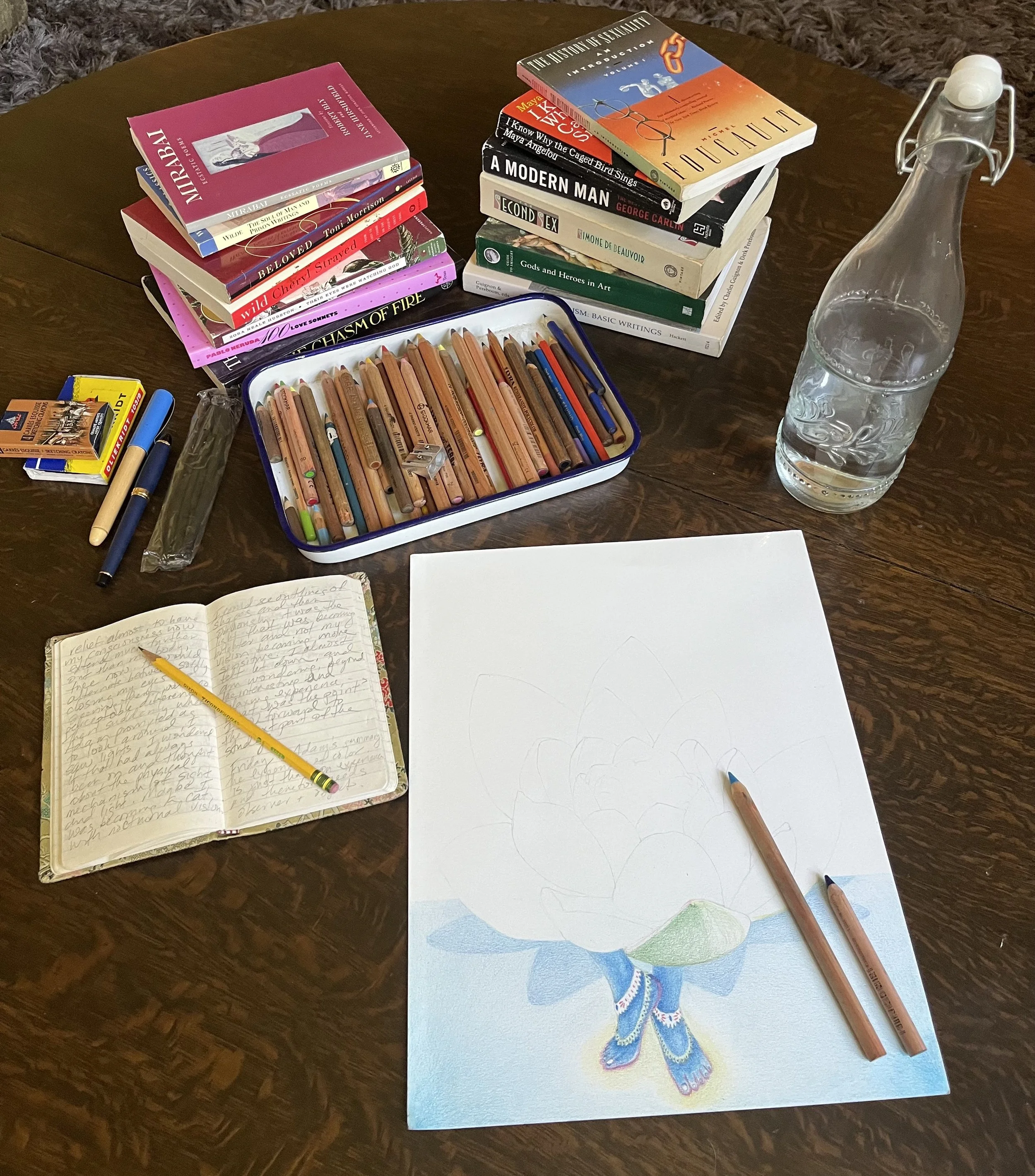 A wooden table with a stack of books, a journal with handwriting, a work of art that is in process, colorful colored pencils, pens, and a glass water bottle.