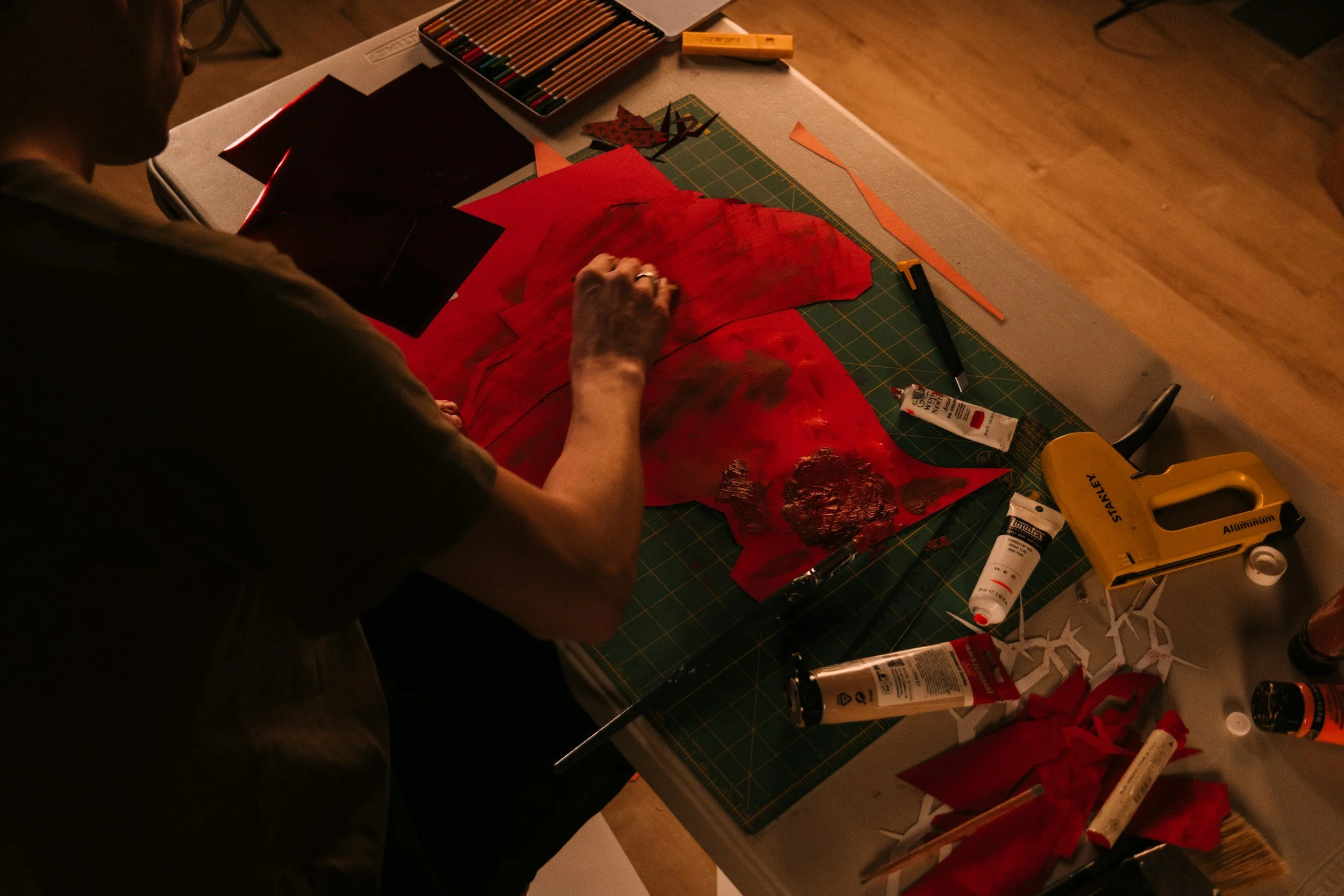 A person working on a craft project at a table with various art supplies, including colored pencils, paper, glue, and tools, in a dimly lit room.