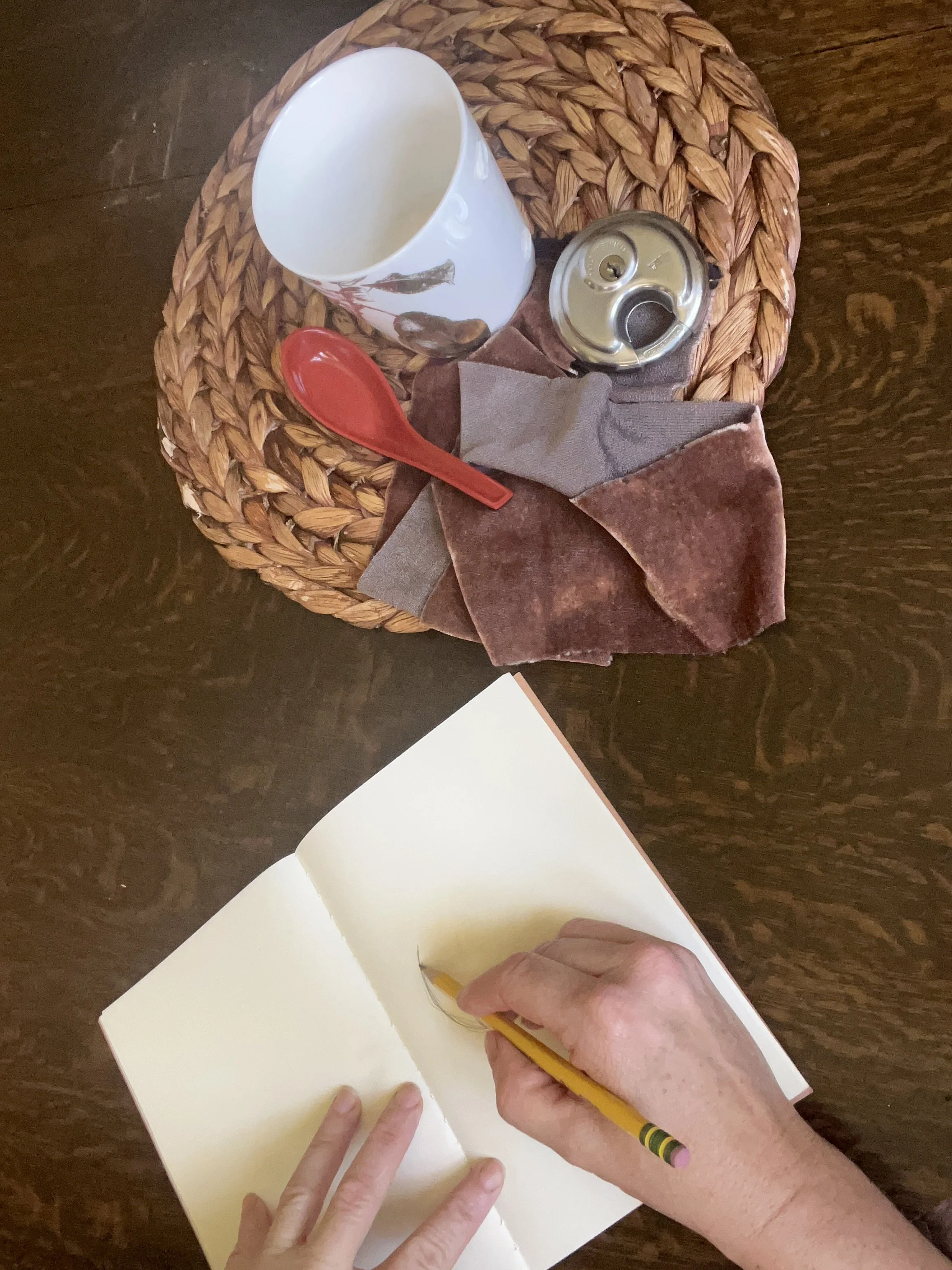 Hands writing in a notebook with a yellow pencil, held in their right hand, on a dark wooden table. Nearby, there is a woven circular placemat with a white mug featuring floral designs, a red Asian soup spoon, a piece of brown vel, and a silver lock.