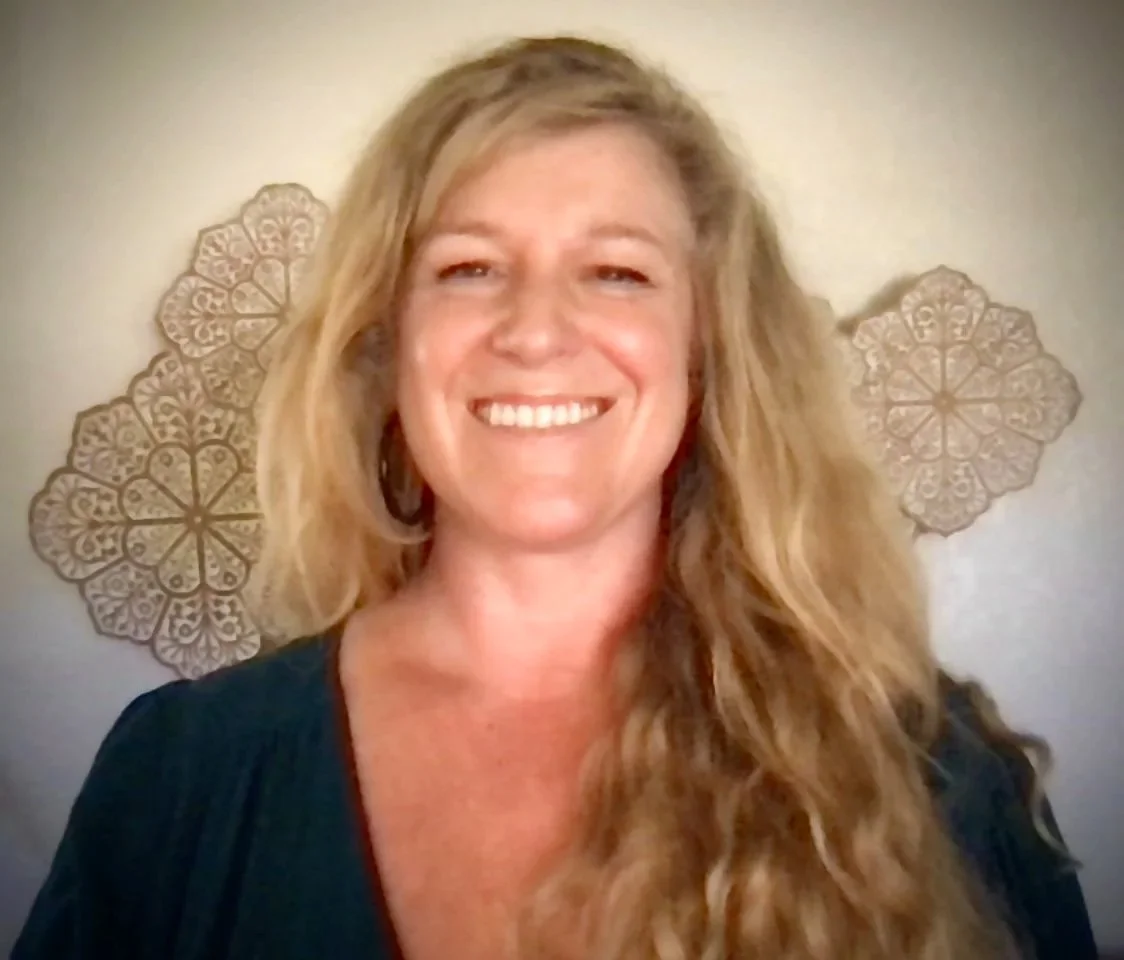 A woman with long, wavy blonde hair smiling in front of a beige wall with decorative floral metal wall art.