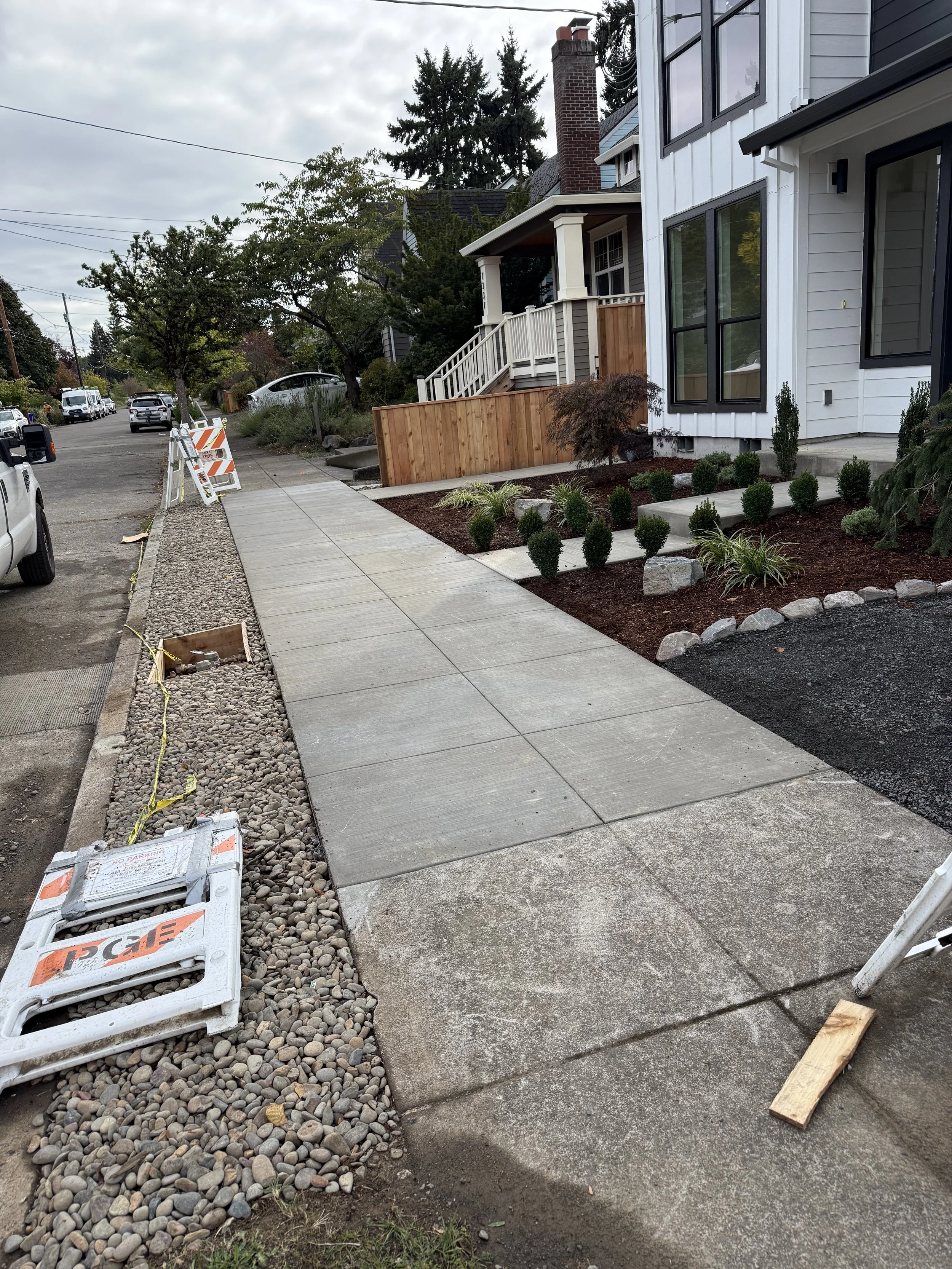 New concrete sidewalk being installed along a residential street, with construction barriers, gravel, and landscaping in progress.