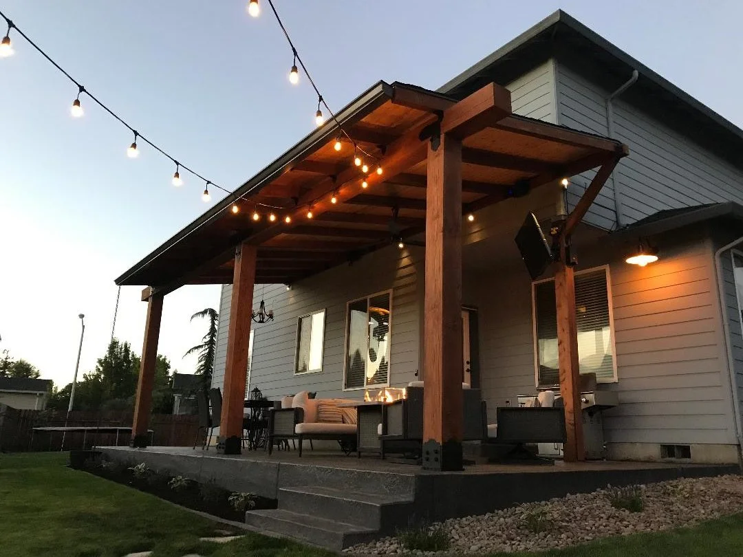 A backyard patio at dusk with string lights overhead, furnished with outdoor seating, a fire pit, and a house with a covered porch.