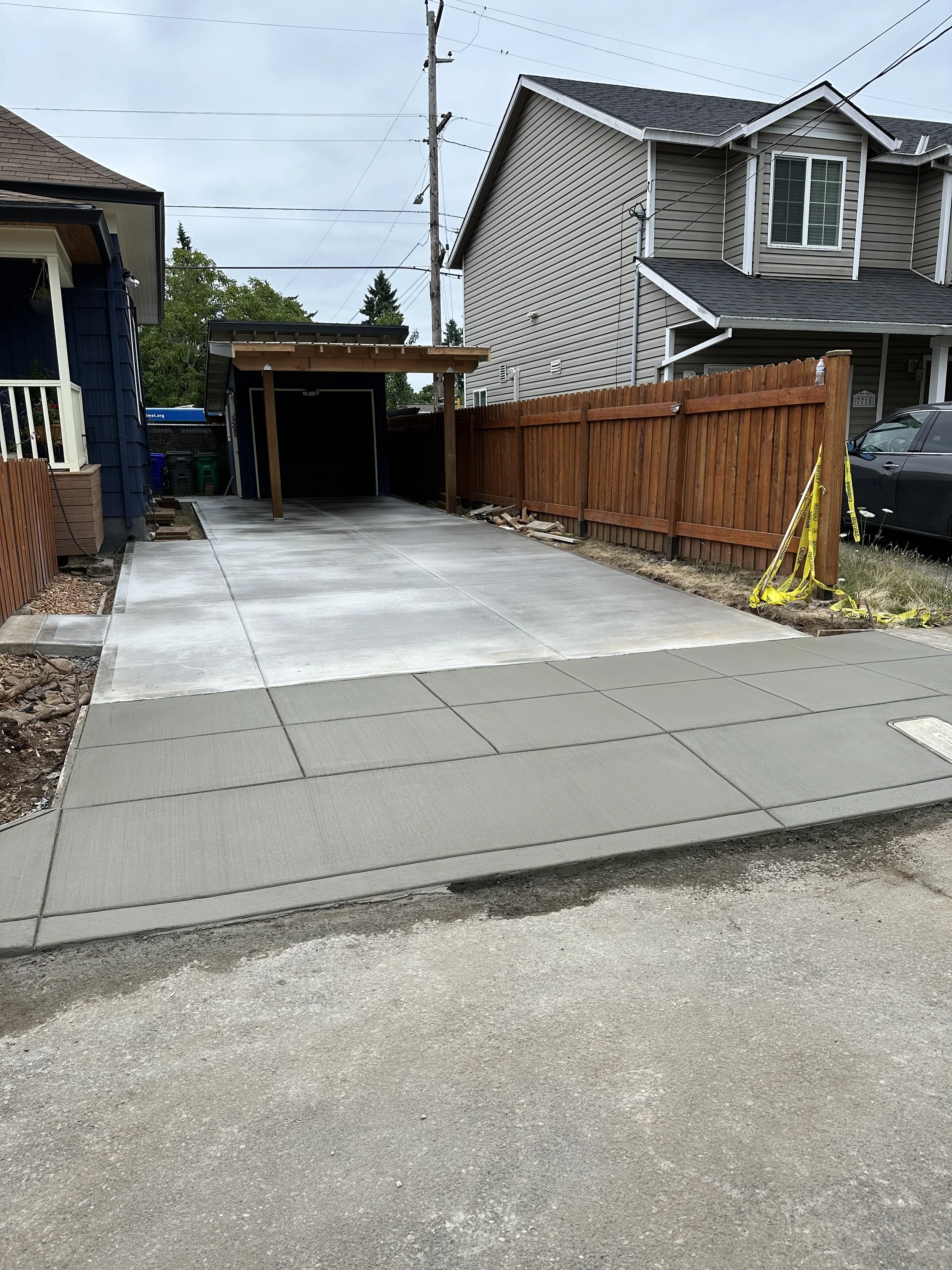 A newly paved concrete driveway in front of a house, with a newly planted wooden fence on the right side and an unfinished sidewalk section at the bottom. There is a car parked beside the house and a small structure with a roof at the end of the driveway.