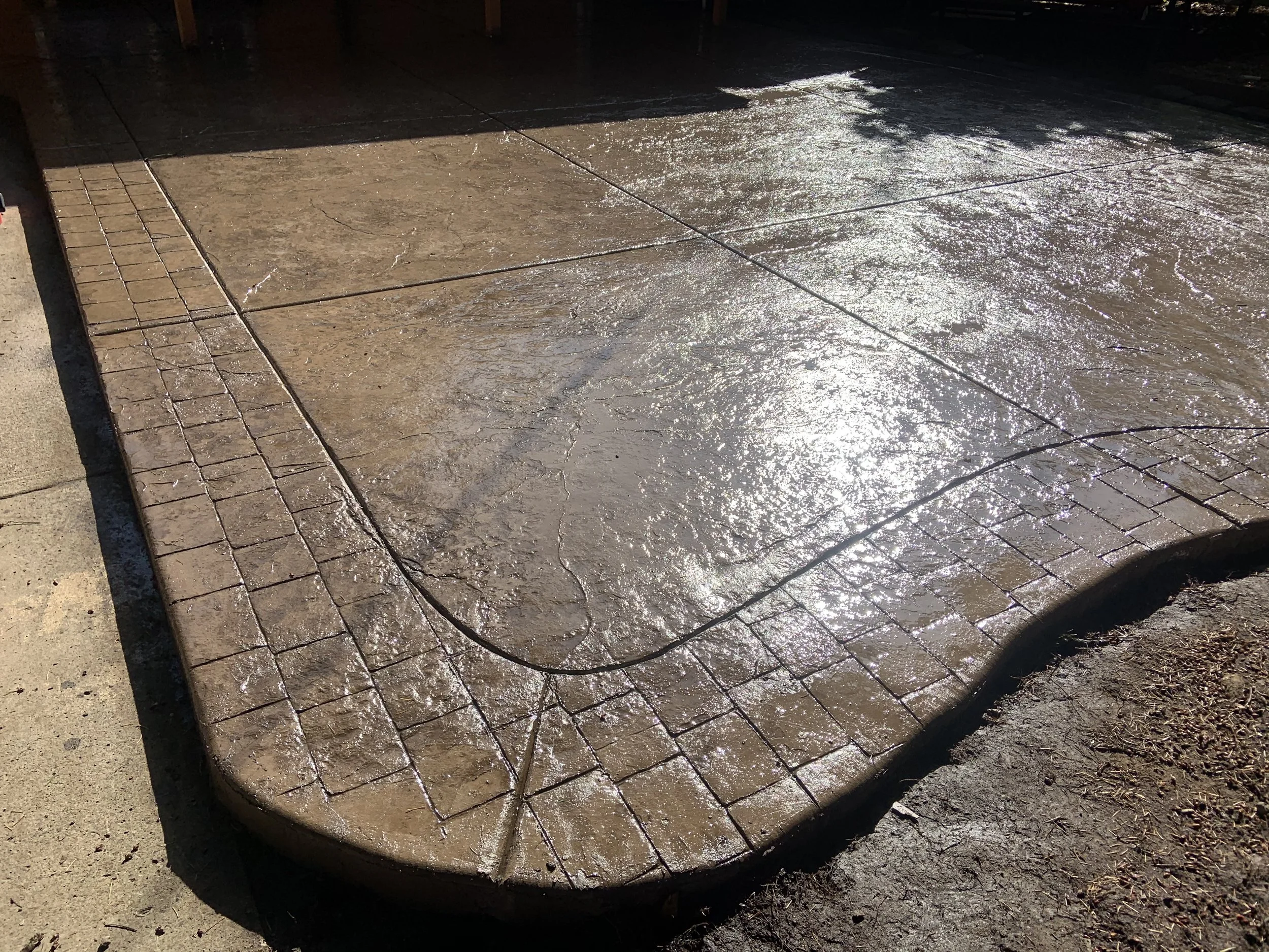 A concrete sidewalk with a stamped brick pattern on the edge, wet and reflecting sunlight.