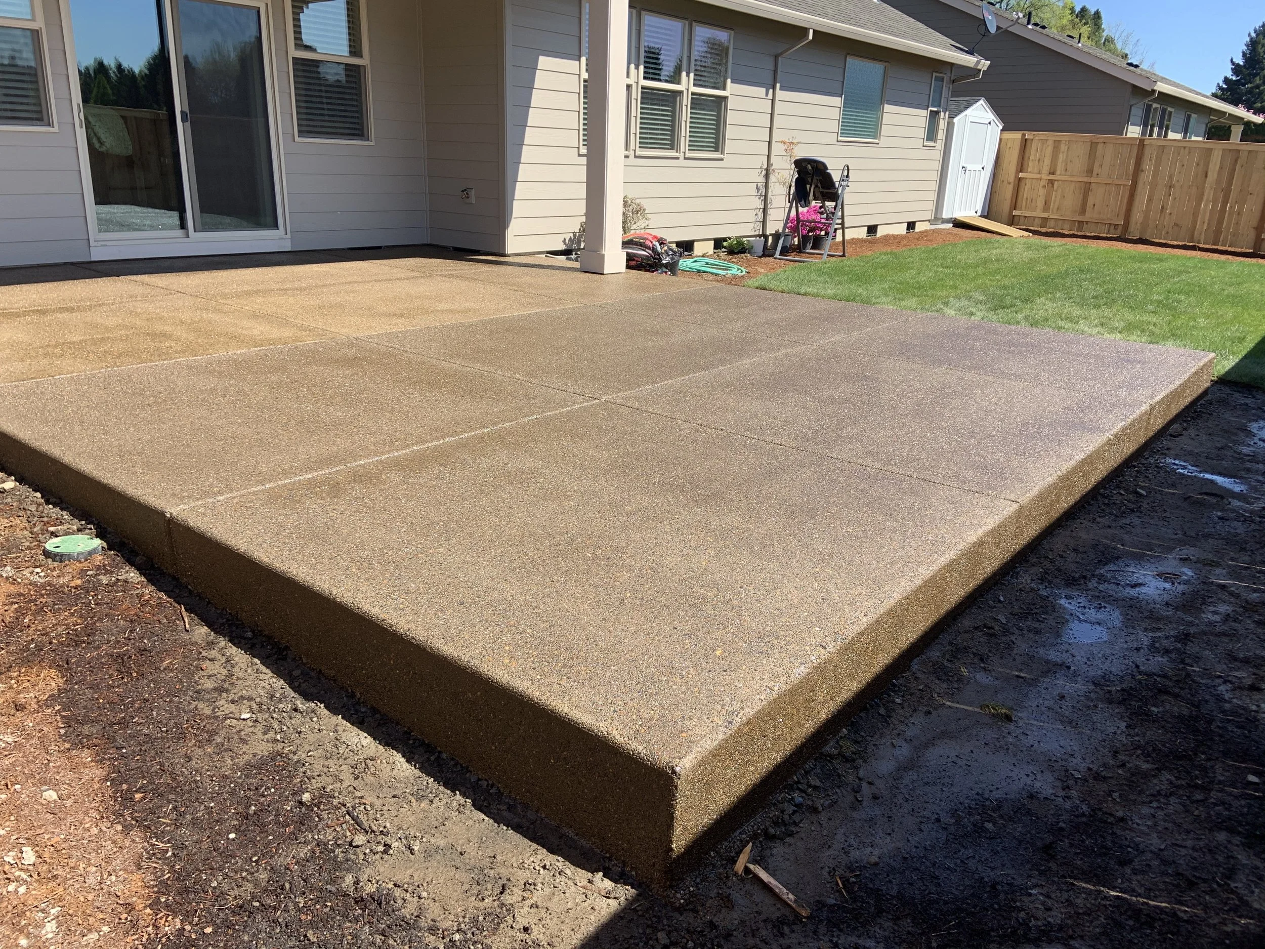 Backyard with freshly poured concrete patio, grass lawn, and a wooden fence in a sunny residential area.