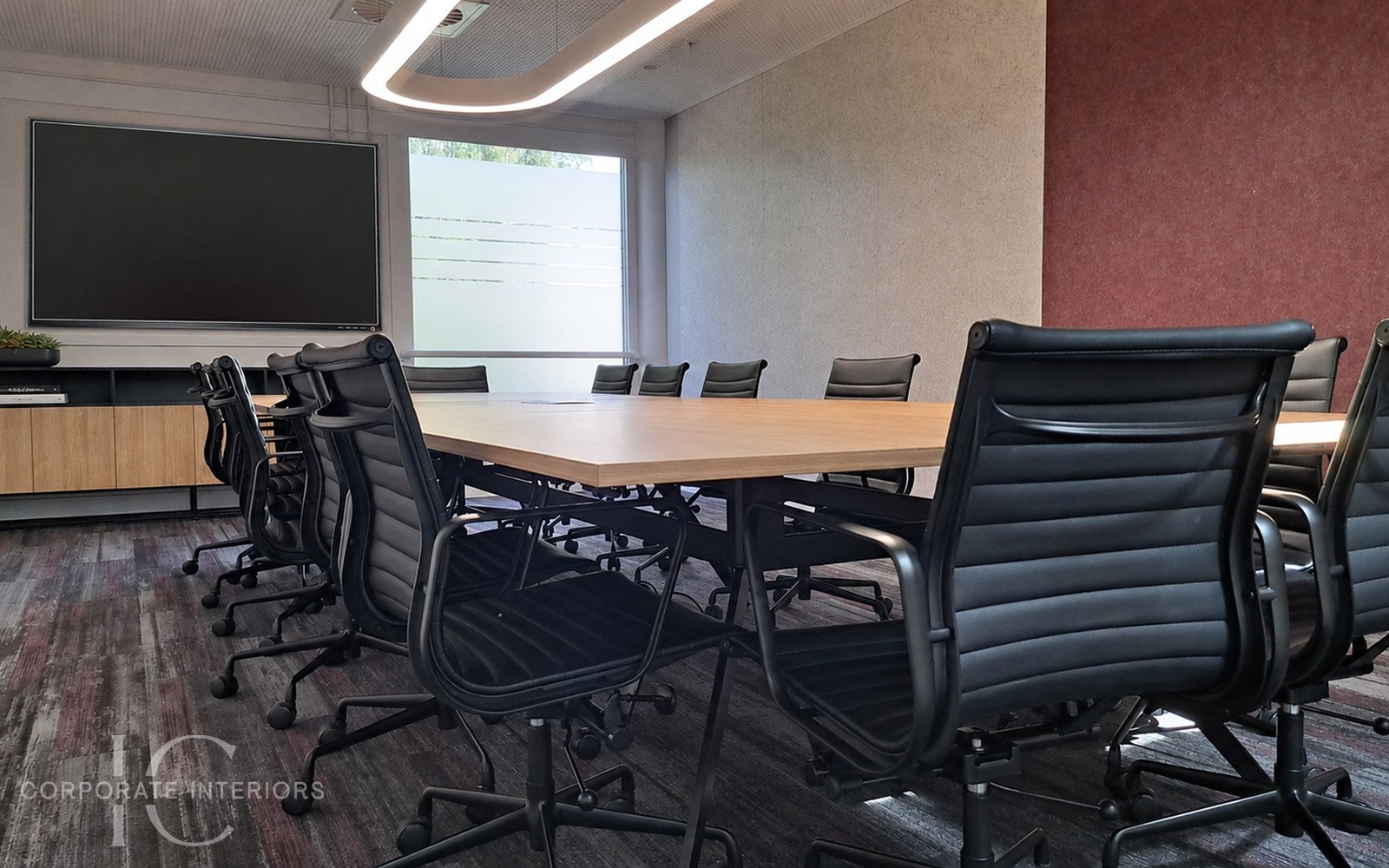 A modern conference room with a long wooden table surrounded by black leather office chairs, a large flat-screen TV mounted on the wall, and a cabinet with decor and books. Natural light enters through large windowed doors.