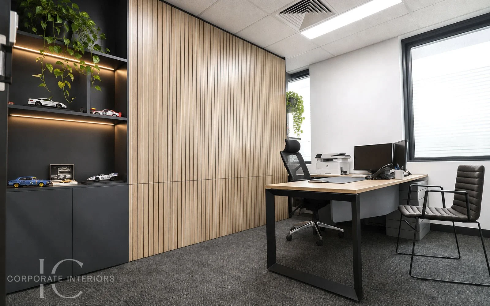 Modern office with a wooden accent wall, black shelving displaying model cars, a desk with computer monitors, a printer, and an office chair, and large windows with city view.