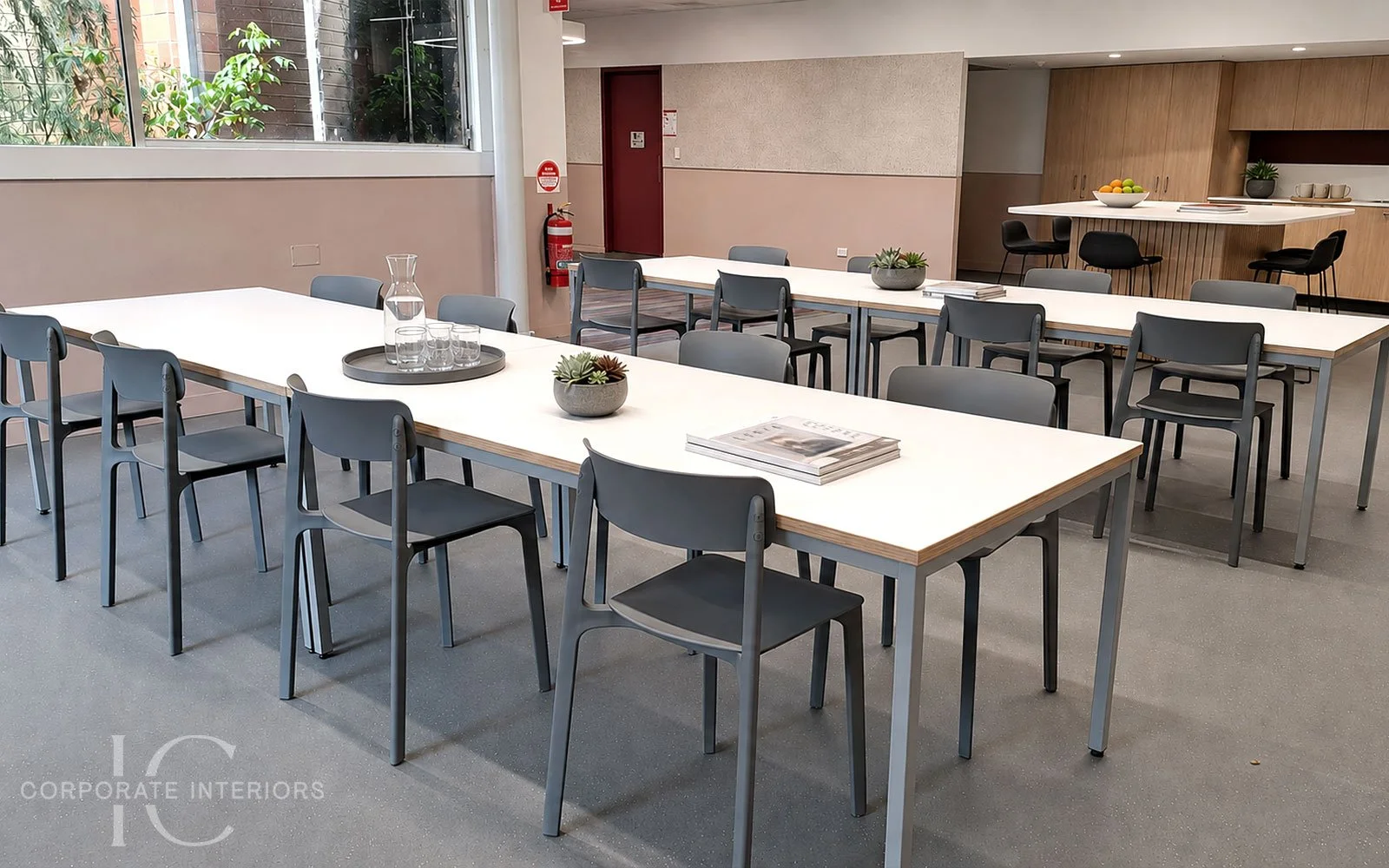Modern conference room with white tables, gray chairs, small potted plants, stacked magazines, and a tray with glass pitcher and glasses. Large window with greenery outside. Wooden cabinet and bowl of fruit in background.