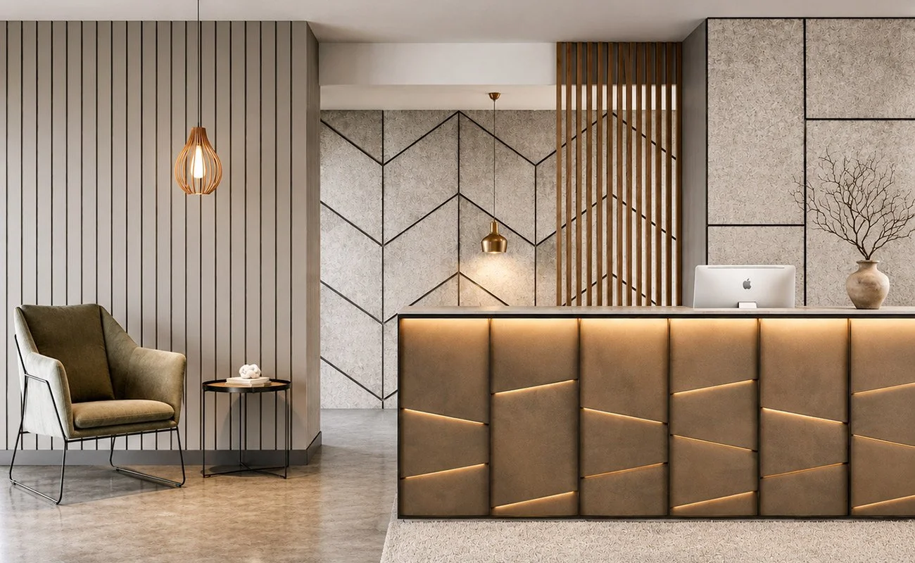 Modern hotel reception area with a beige textured front desk, a desktop computer, and decorative branches in a vase. To the left, there is a beige and dark green armchair beside a small black side table with a rolled-up towel or cloth. Two pendant lights, one copper and one gold, hang from the ceiling. The background features textured wall panels with geometric patterns and wooden slats.