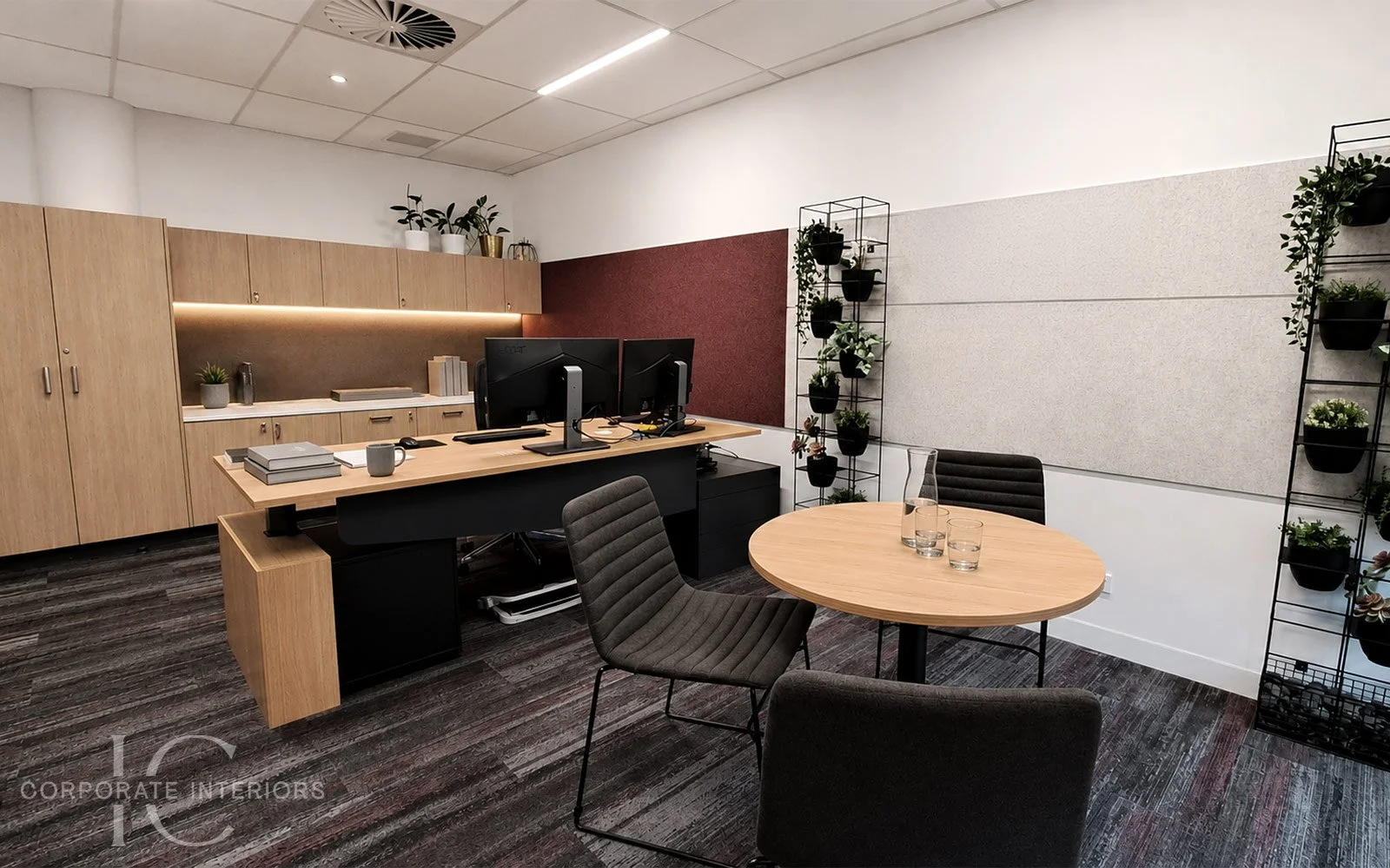 Modern office with wooden cabinets, dual computer monitors on a desk, table with chairs, and decorative plant shelving.