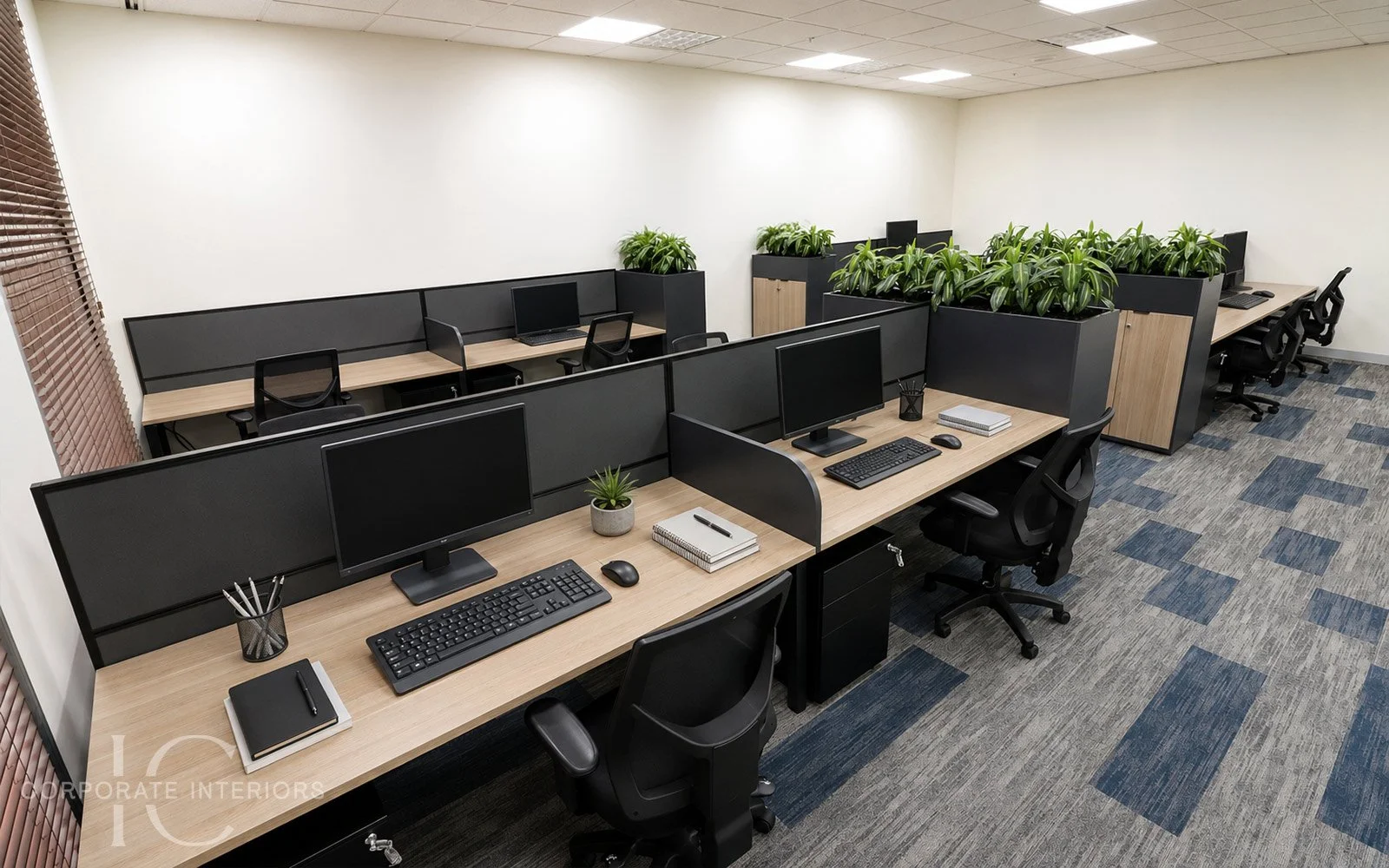 Modern office with multiple workstations separated by black dividers, each equipped with a computer, monitor, keyboard, mouse, and office supplies. Large green plants in black planters add a touch of nature. The floor features a patterned carpet, and