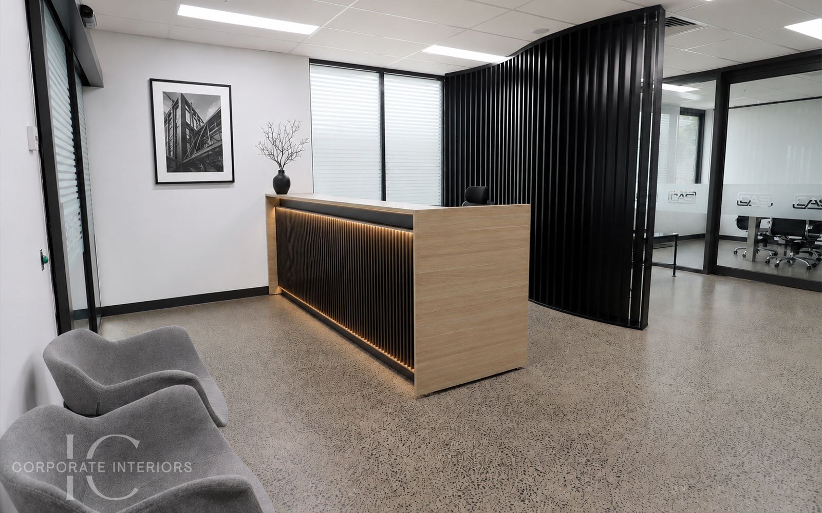 Modern office reception area with a curved wooden and black slatted reception desk, gray chairs, black artwork, and a glass-walled meeting room in the background.