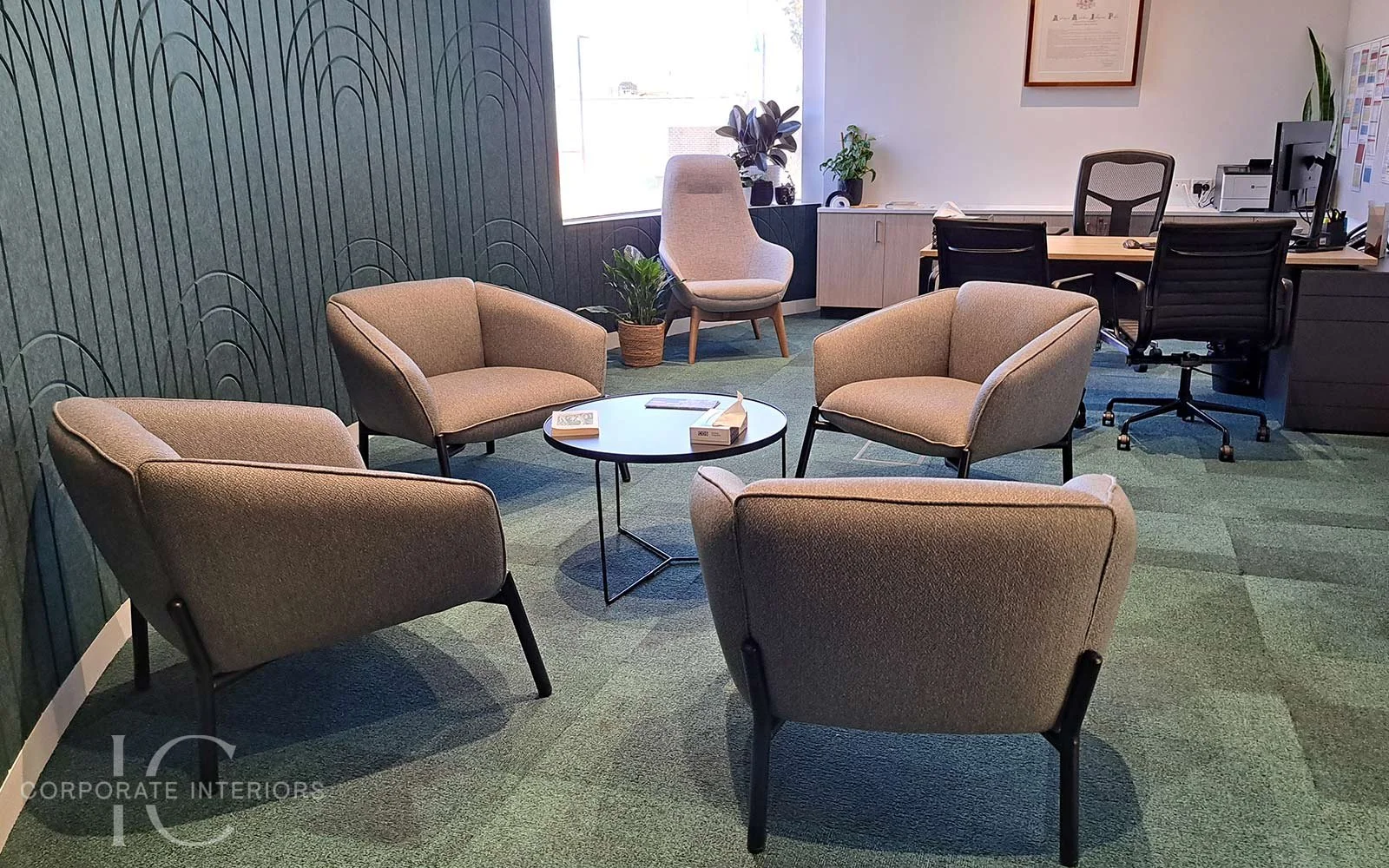 An office waiting area with five beige armchairs arranged around a round black coffee table, a potted plant, a sitting area with a high-back chair near a window, and a corporate desk with a computer and office supplies in the background.