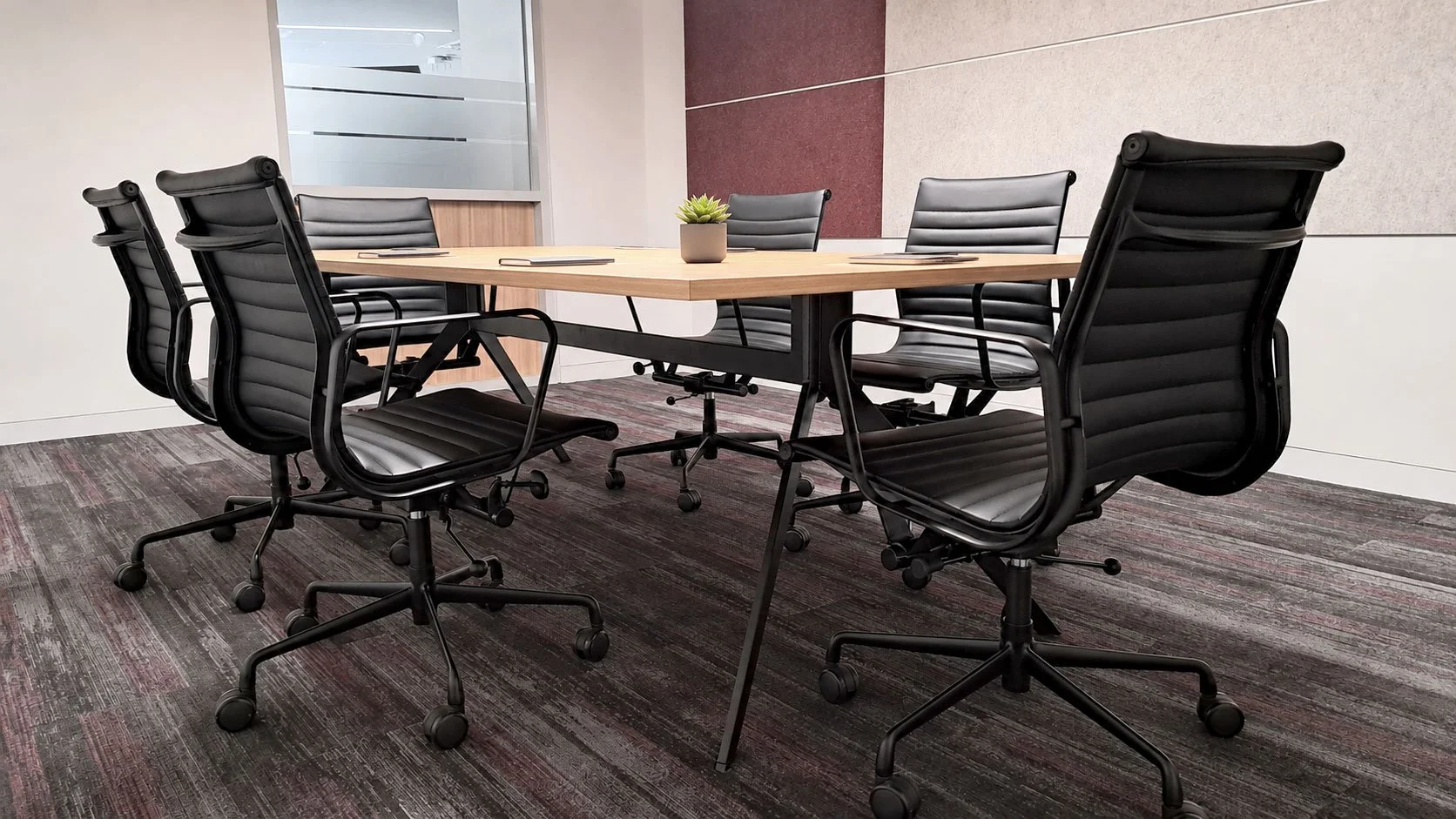 An empty modern conference room with a rectangular wooden table and six black leather office chairs around it, a small potted plant in the center of the table, carpeted flooring, and a glass wall in the background.