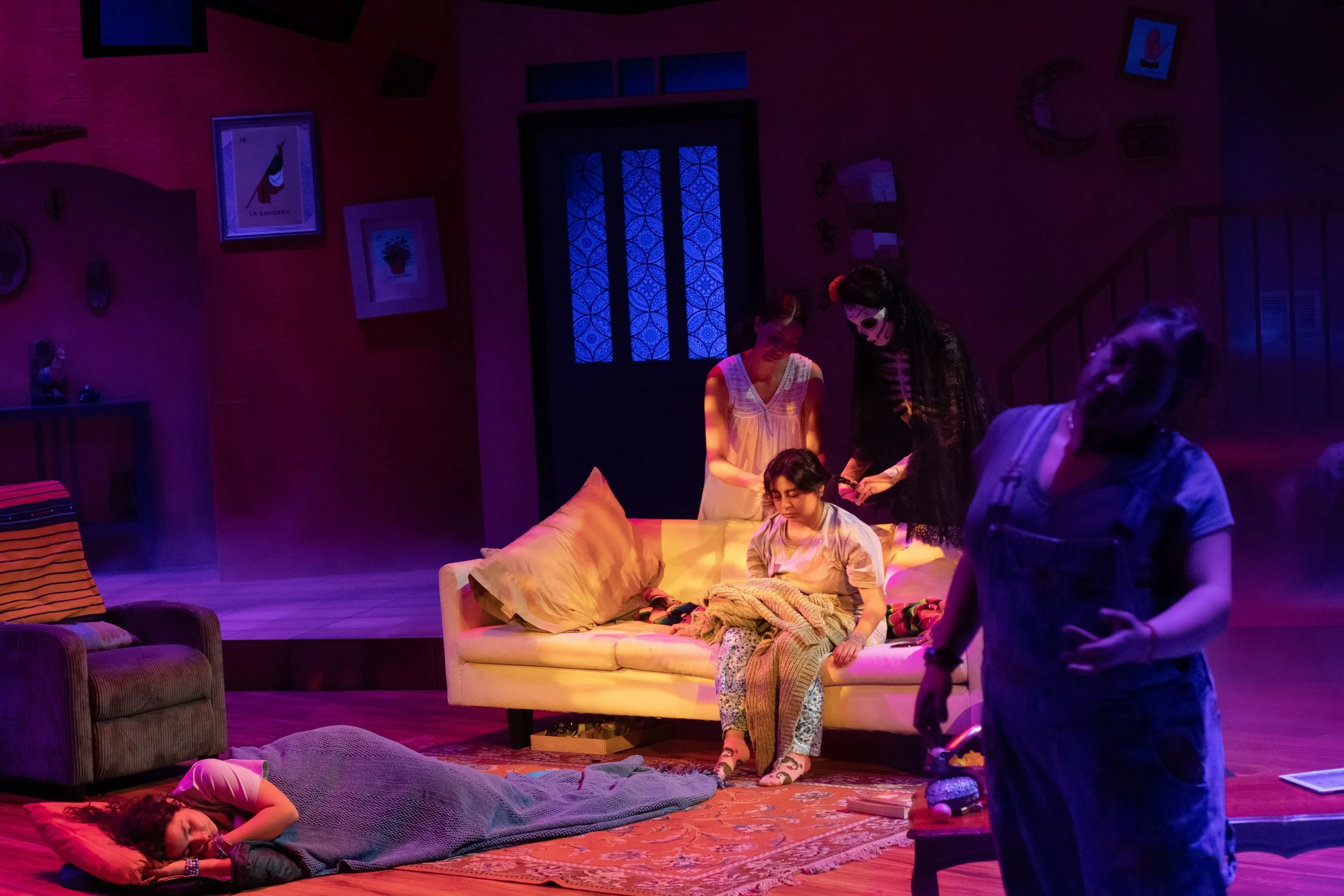 Scene from a theatrical play with children playing dead on the floor, sitting on a couch, and interacting in a cozy, dimly lit room with colorful lighting, sofas, framed pictures, and a door with a decorative glass panel.