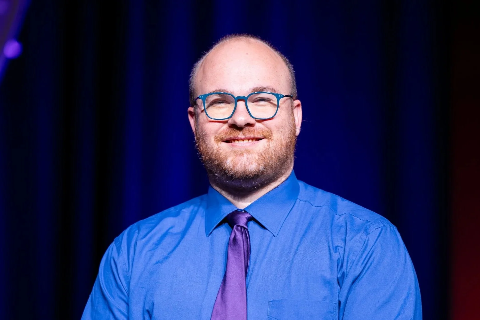 Smiling man wearing blue glasses, blue dress shirt, and purple tie against a dark blue background