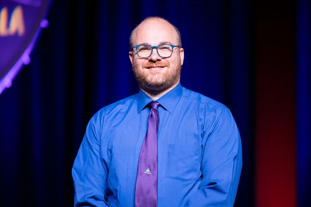 A man with glasses, a beard, and short hair wearing a blue dress shirt and a purple tie, standing on a stage with a dark background.